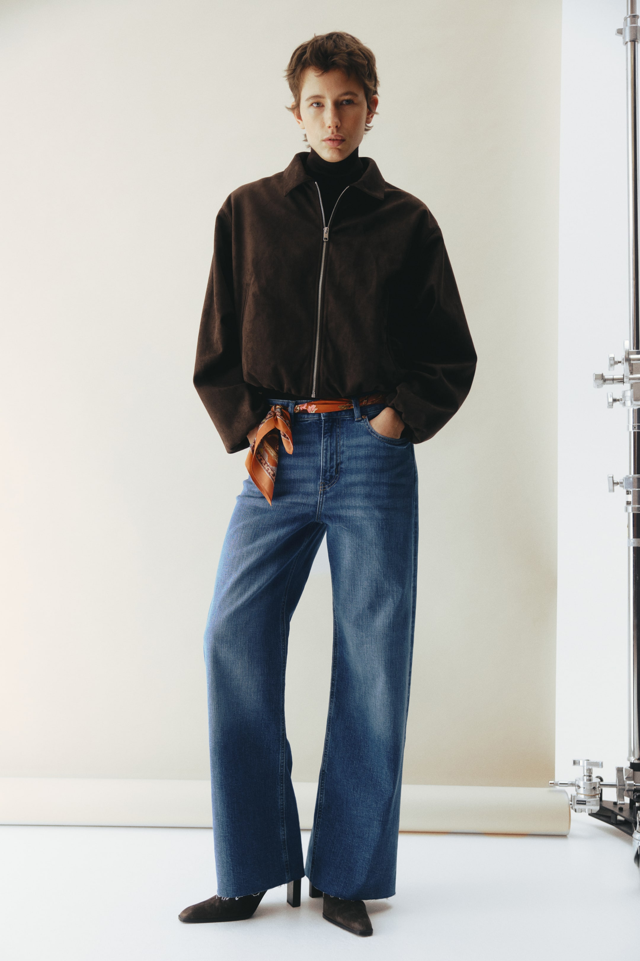 View larger image: A dark brown suede-like jacket features a silver-tone full-zip closure and a pointed collar, worn over a black turtleneck. It is paired with high-waisted, wide-leg blue jeans with a raw hem and a small patterned scarf tied as a belt.