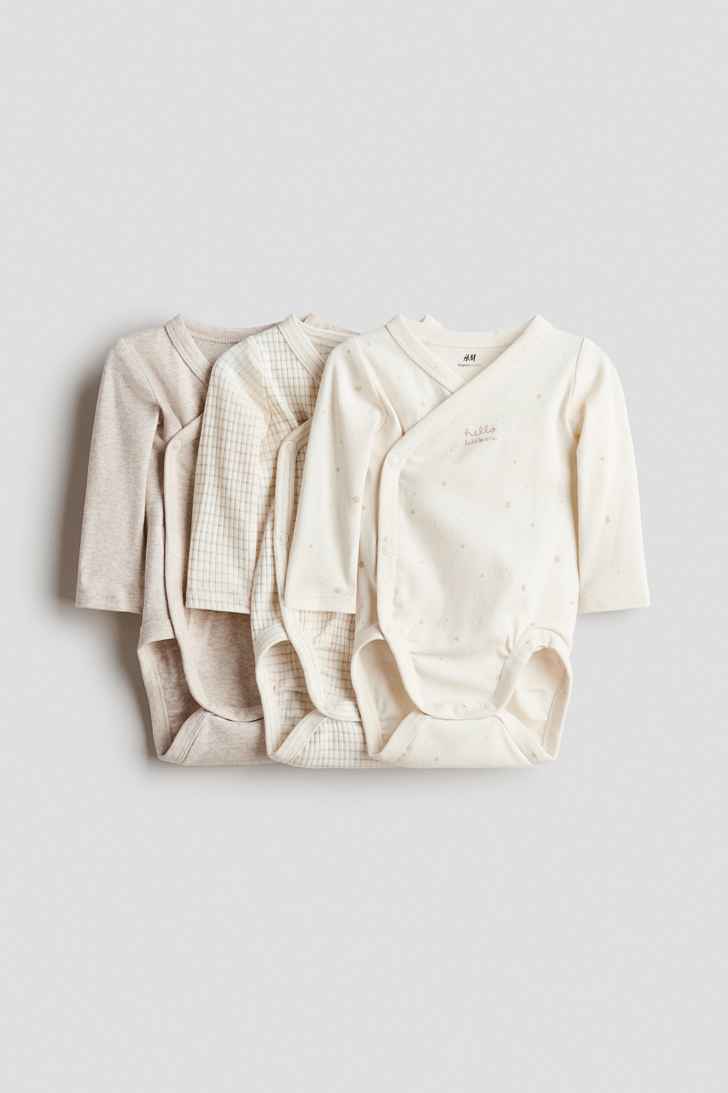 A long-sleeved baby bodysuit in a cream grid pattern is shown from the front, featuring a wrap-front design with bound trim. The bodysuit is being worn by an infant lying down, who is also styled with a matching cream knit bonnet and booties.