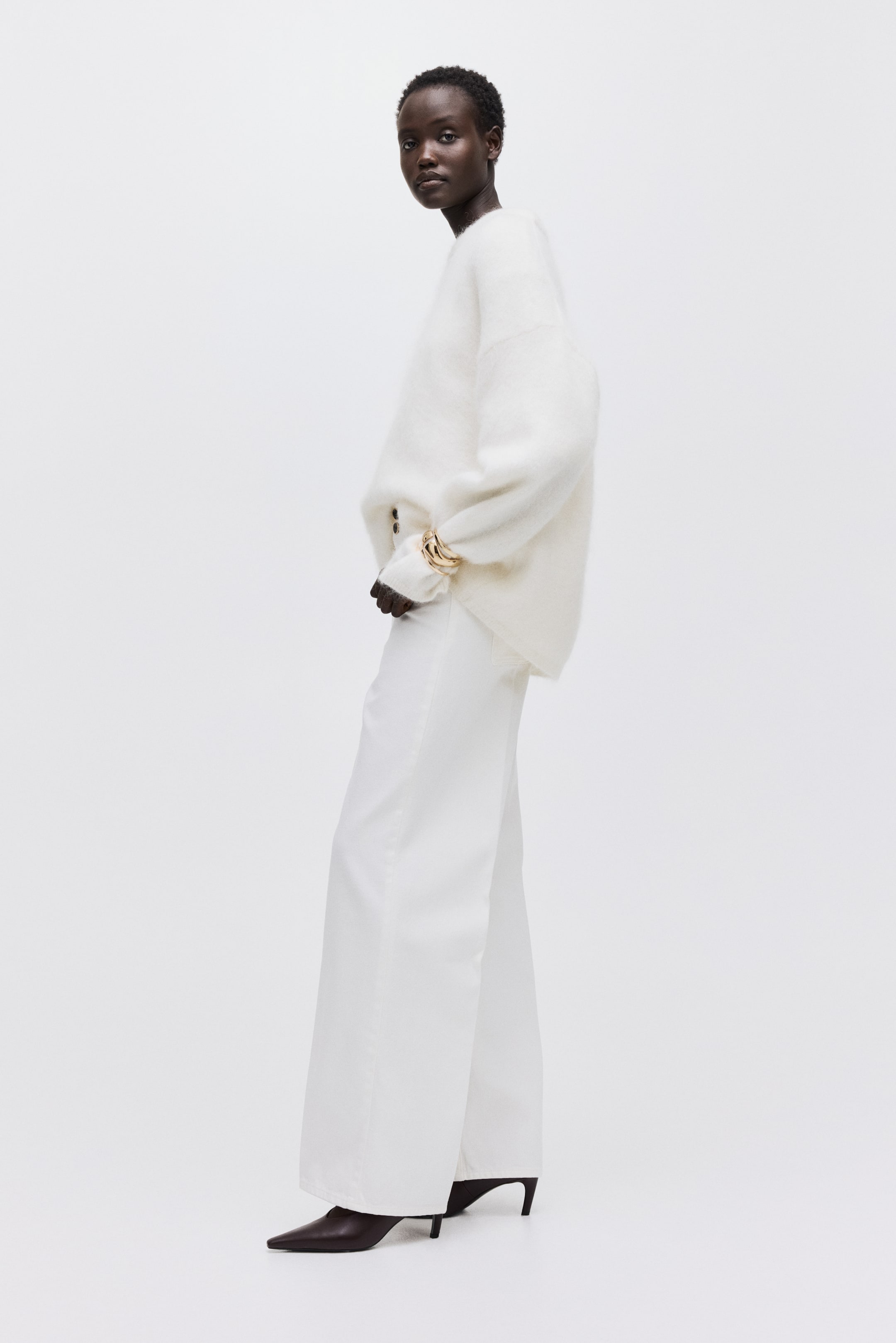 Voir une image plus grande: Un pull blanc duveteux et surdimensionné est présenté de biais, avec un col rond et de longues manches amples. Il est assorti à un pantalon blanc à jambes larges qui tombe sur des chaussures pointues foncées, avec un bracelet doré visible sur une manche du pull.
