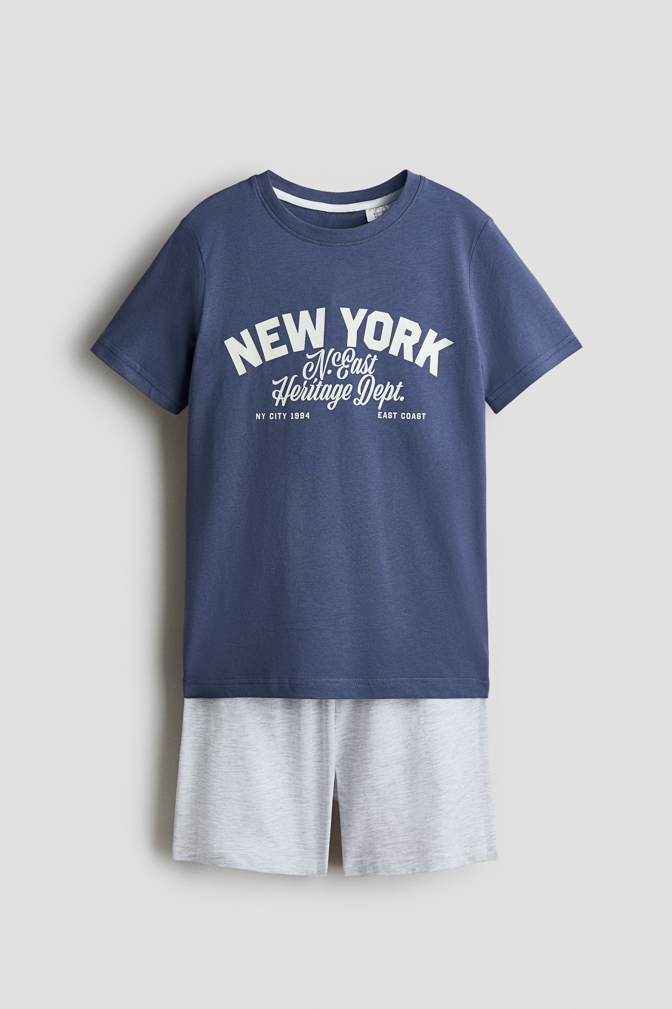 Prikaži veću sliku: Prikazan je komplet pidžame sa tamnoplavom T-majicom na čijem se prednjem delu nalazi bela grafika 'NEW YORK N.East Heritage Dept. NY CITY 1984 EAST COAST' i rebrastim okruglim izrezom oko vrata. Usklađene svetlosive kratke hlače imaju elastični pojas u struku.