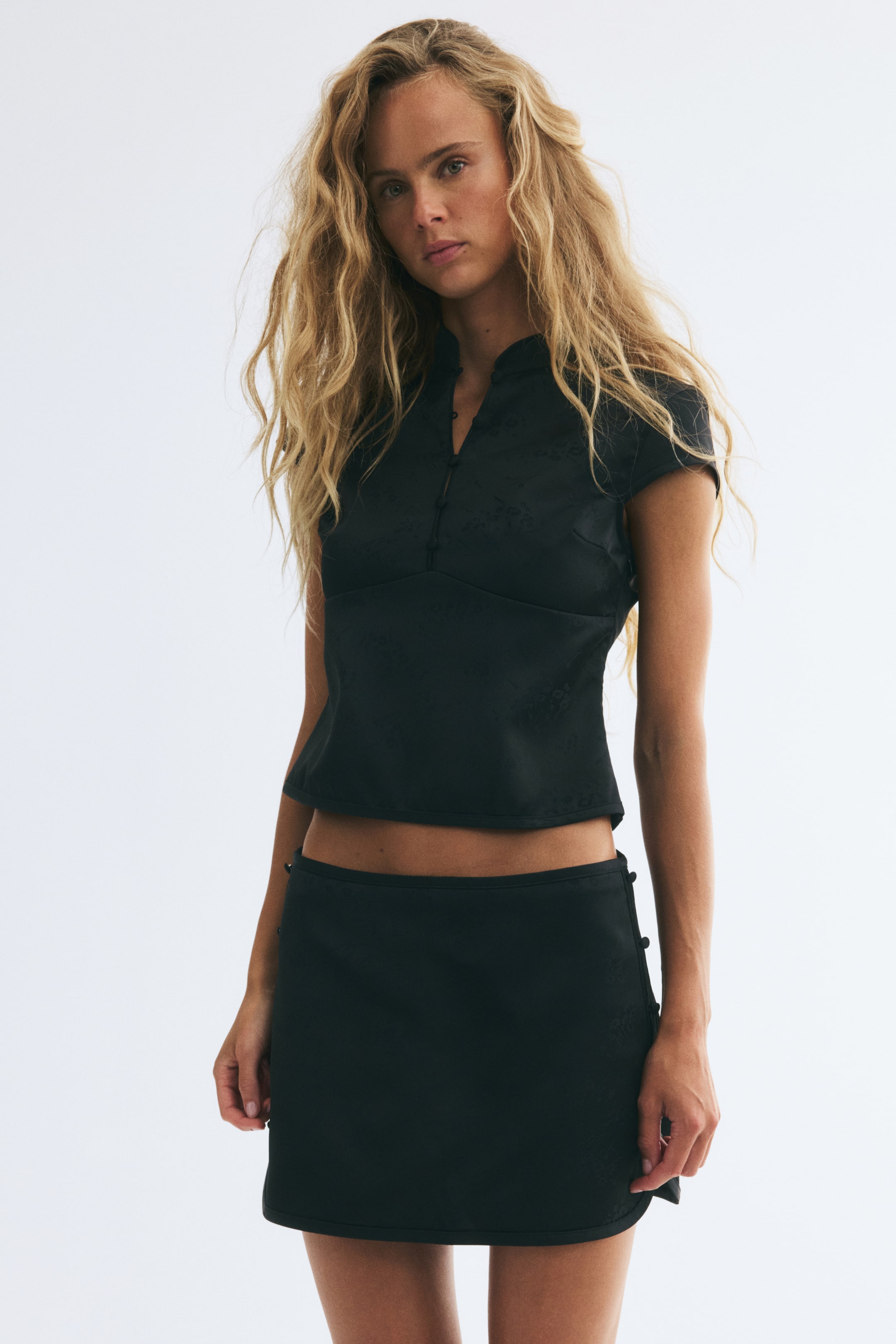 View larger image: The model is wearing a black mini skort in textured jacquard fabric, featuring a low waist and small decorative buttons at the sides. The skort is styled with a matching black short-sleeved top.