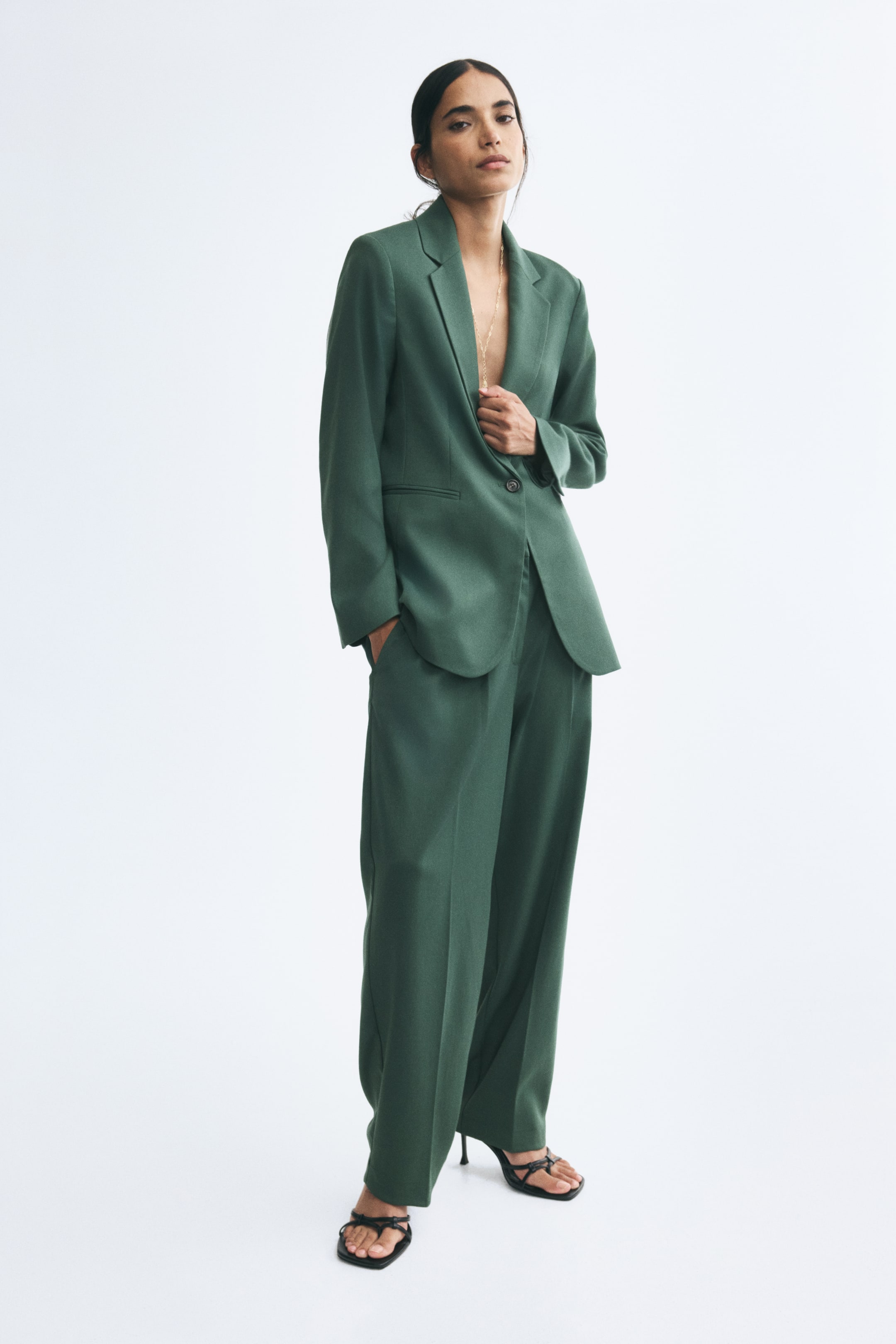 View larger image: Woven green pants with straight legs and front creases are worn by the model, who is standing with one hand resting in the side pocket. The pants are styled with a matching green blazer and black strappy heeled sandals.