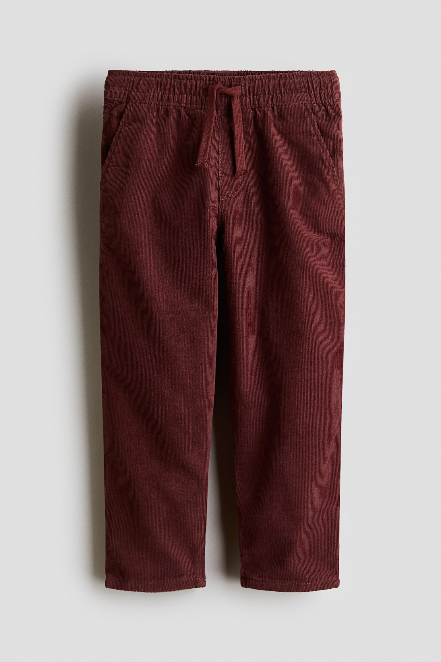 A pair of burgundy cotton corduroy trousers is shown from the front, featuring an elasticated waistband with a matching drawstring and a mock fly. The design showcases a loose fit through the hips and thighs, with gently tapered legs and side pockets.