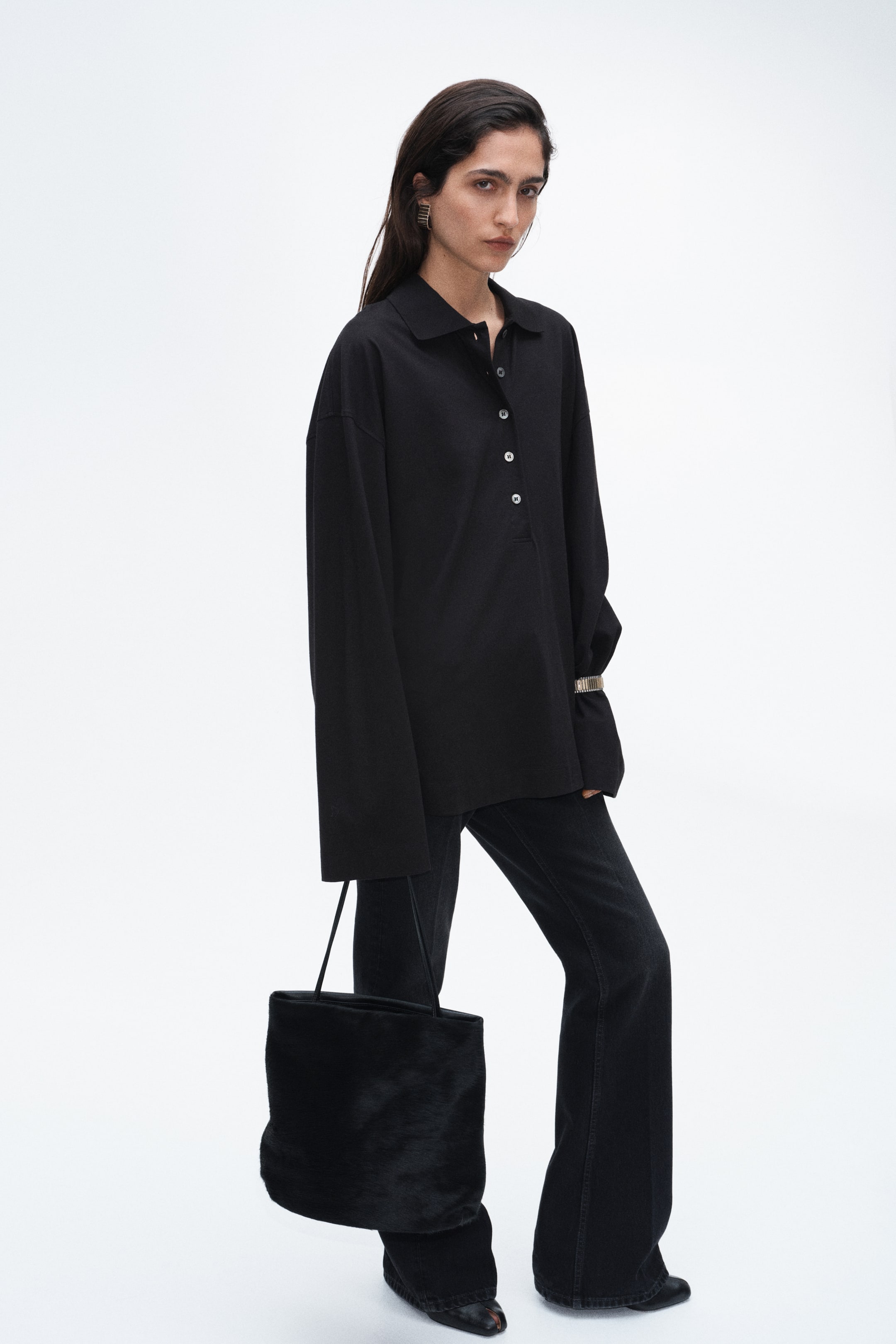Se større billede: En sort oversized poloshirt med krave og knaplukning bæres af modellen. Modellen har også sorte flared jeans på og bærer en sort tote-taske i imiteret pels. Et cuffarmbånd i guldfarvet finish bæres uden på skjortens ærme.