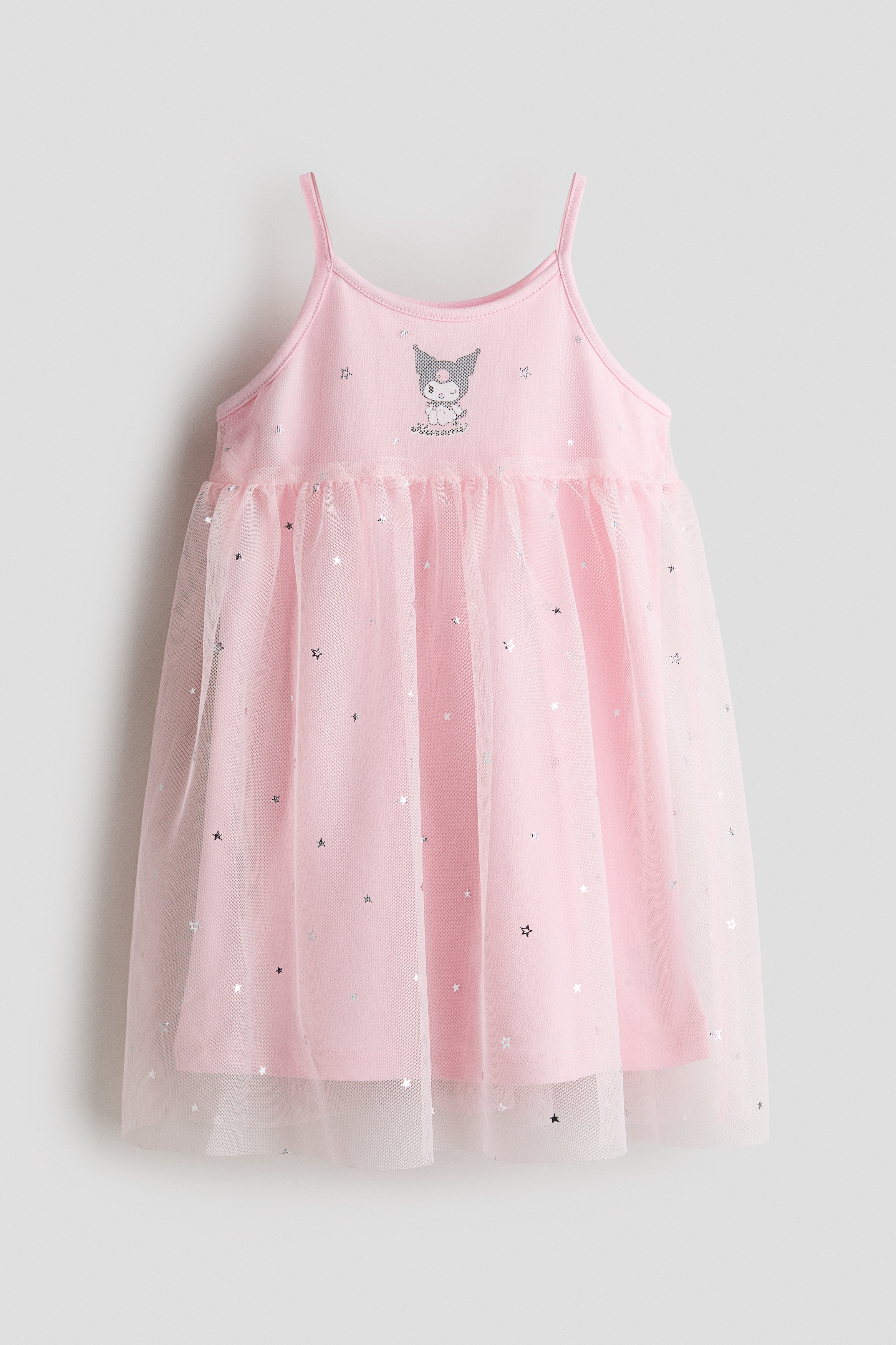 A pink sleeveless dress with slim shoulder straps and a round neckline is being worn by the model. The dress features a character graphic on the chest and a flared skirt with a sheer tulle overlay decorated with small silver stars, which the model is holding gently at the side.