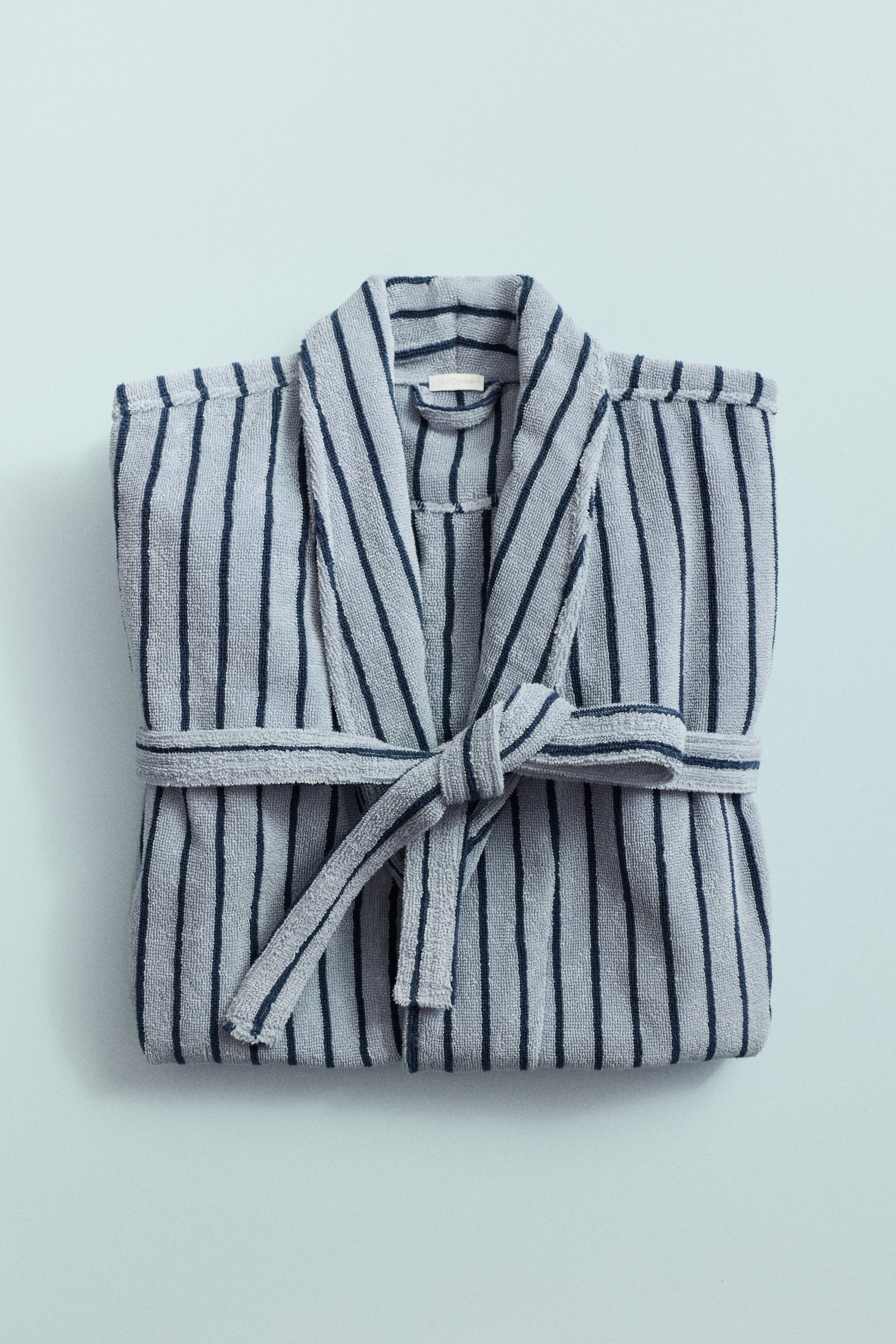 View larger image: A terry cloth bathrobe is presented folded flat, featuring light blue-gray and dark blue vertical stripes. It is designed with a shawl collar, and a matching tie belt is knotted at its front.
