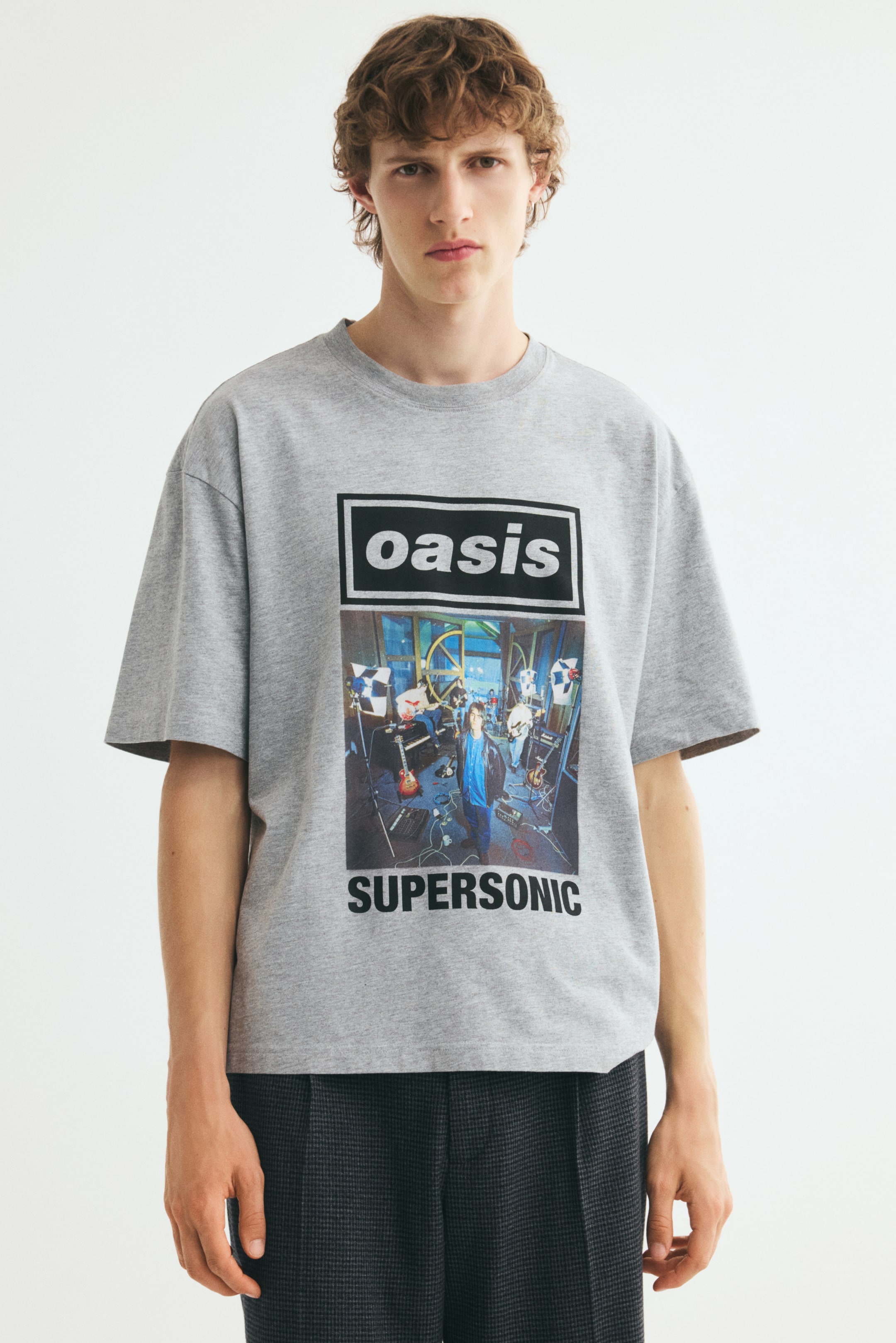 Ver imagen más grande: La modelo lleva una camiseta de punto gris de corte relajado con cuello redondo acanalado, hombros caídos y un estampado gráfico frontal con las palabras "oasis" y "SUPERSONIC". La camiseta se combina con un pantalón oscuro de textura marcada.