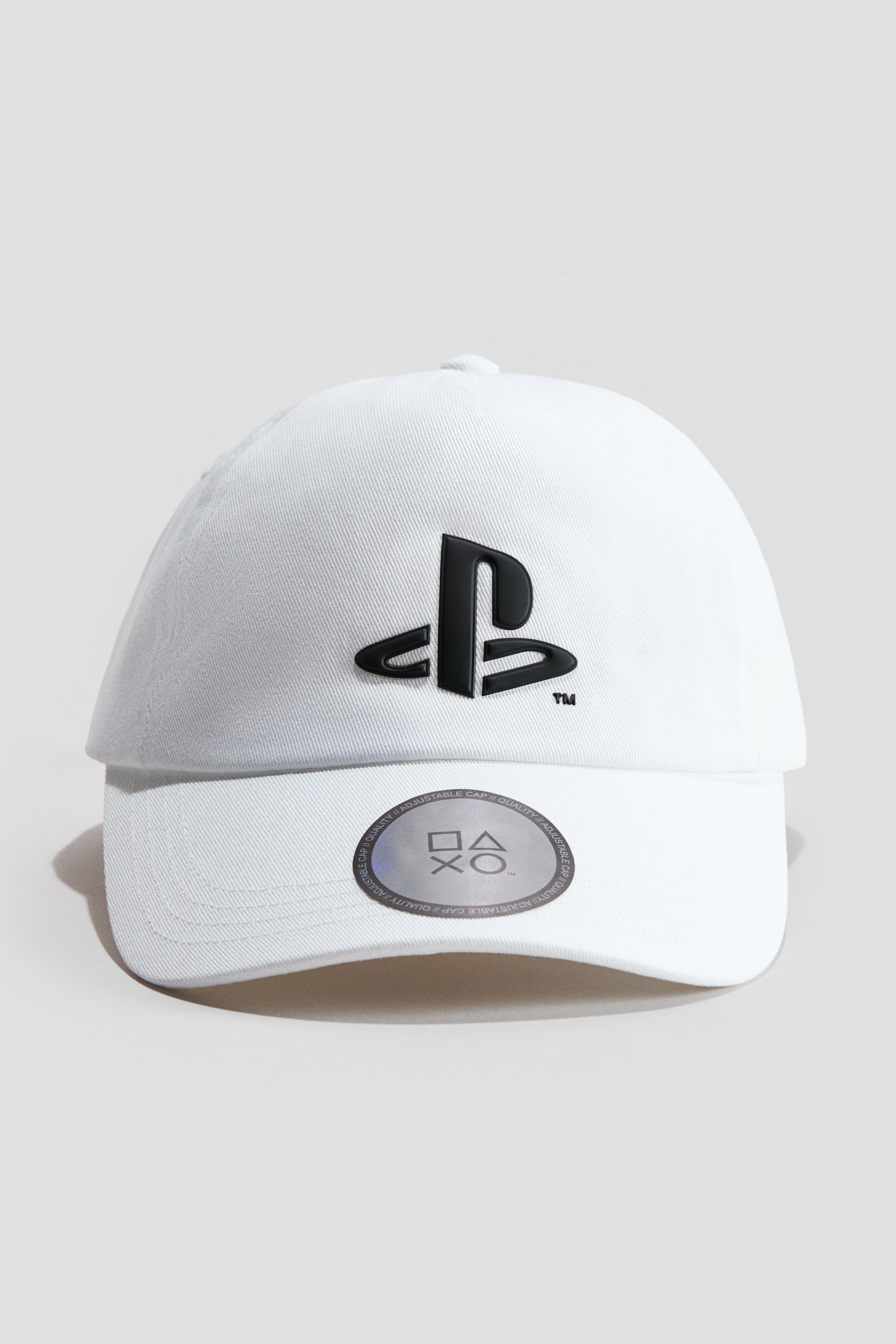 Ver imagen más grande: Una gorra de sarga de algodón blanco está orientada hacia el frente, mostrando un logo de PlayStation en relieve negro en su corona. Un adhesivo circular plateado con símbolos de controladores de PlayStation está adherido a la visera curva.