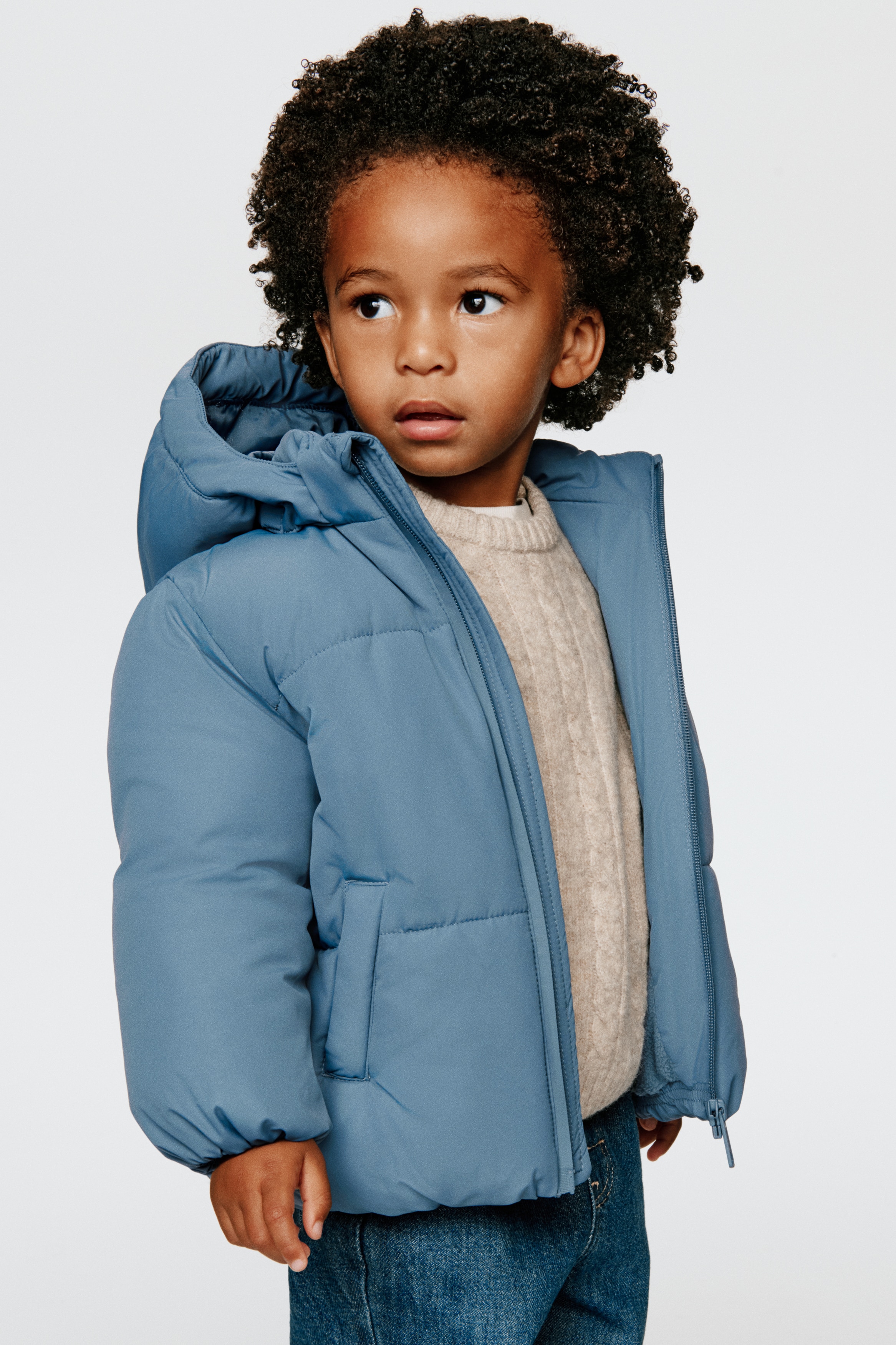 A padded jacket in a dusty blue colour is shown from the front, featuring a lined hood, a stand-up collar, and a full-length front zipper with a chin guard. The jacket features horizontal quilting, vertical welt pockets, and gathered, elasticated cuffs and hem.