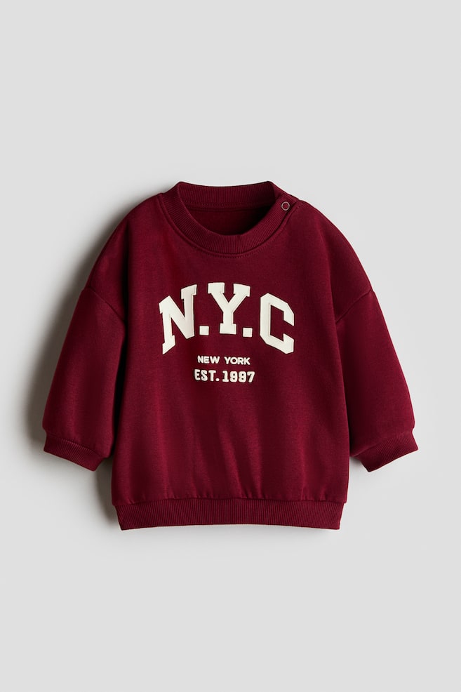 The maroon printed sweatshirt is displayed from the front, showcasing a prominent white "N.Y.C" graphic, accompanied by "NEW YORK" and "EST. 1997" printed below. It features a ribbed crew neck with two snap buttons on the left shoulder, along with ribbed cuffs and a ribbed hem. - 1