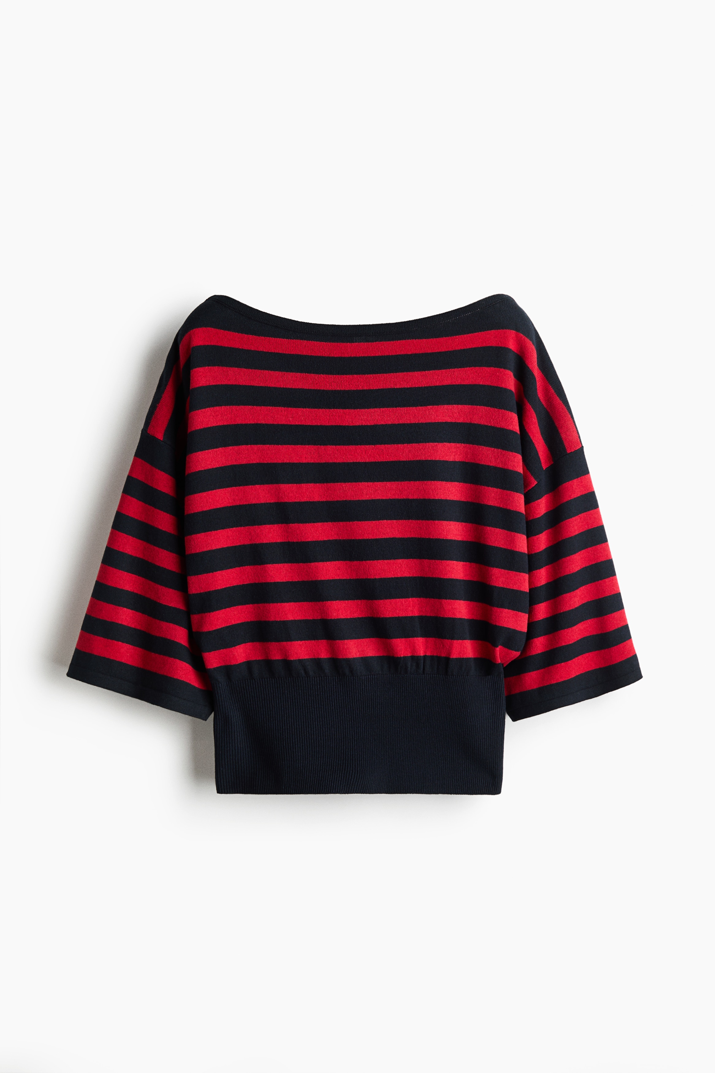 A modelo está a usar uma camisola de malha às riscas horizontais vermelhas e pretas, com decote barco, mangas a meio do braço e uma cinta larga preta ajustada à cintura. A camisola é combinada com calças pretas de perna larga, enquanto a modelo posa com as mãos levantadas atrás da cabeça.