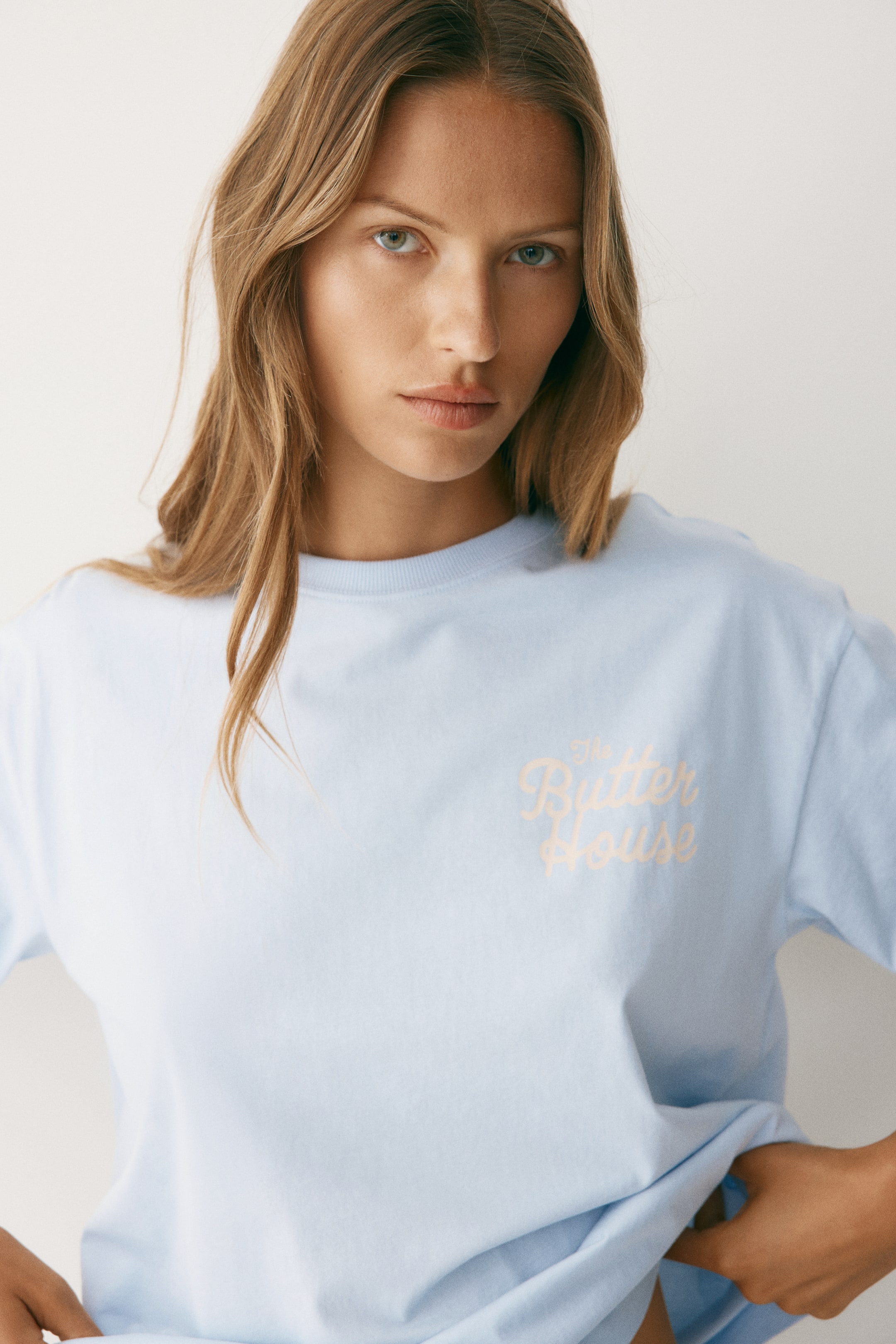 View larger image: A light blue t-shirt featuring a round neck and short sleeves, with "The Butter House" printed in a light peach script font on the upper right chest. The bottom hem of the relaxed-fit shirt is held by hands, emphasizing its loose structure.