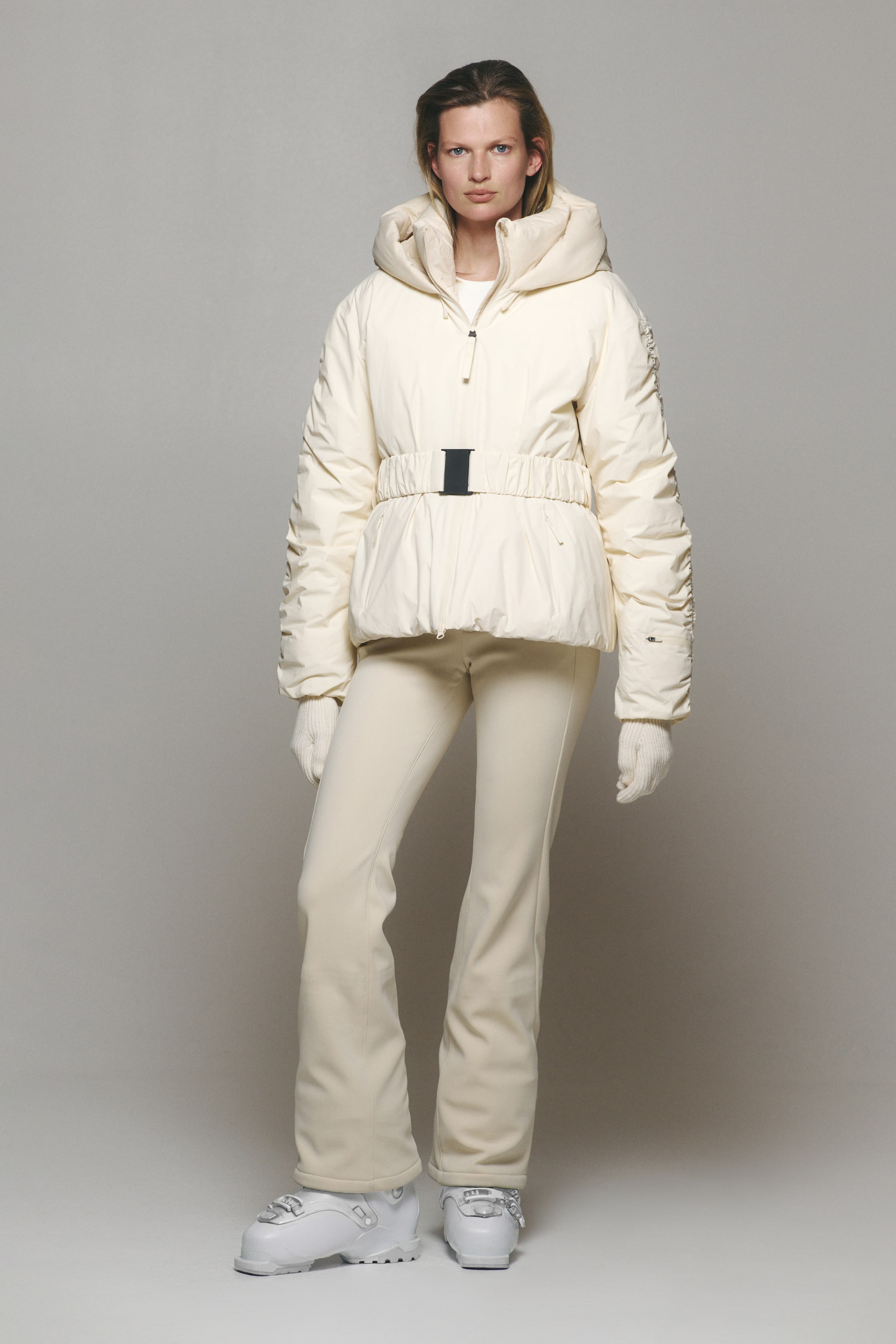 Agrandir l'image: Le mannequin porte une veste de ski matelassée écru, ceinturée, avec un col montant à capuche et une ceinture à la taille élastiquée ornée d’une boucle noire contrastante. La veste est associée à un pantalon de ski slim assorti, des gants en maille blancs et des bottes de ski blanches.