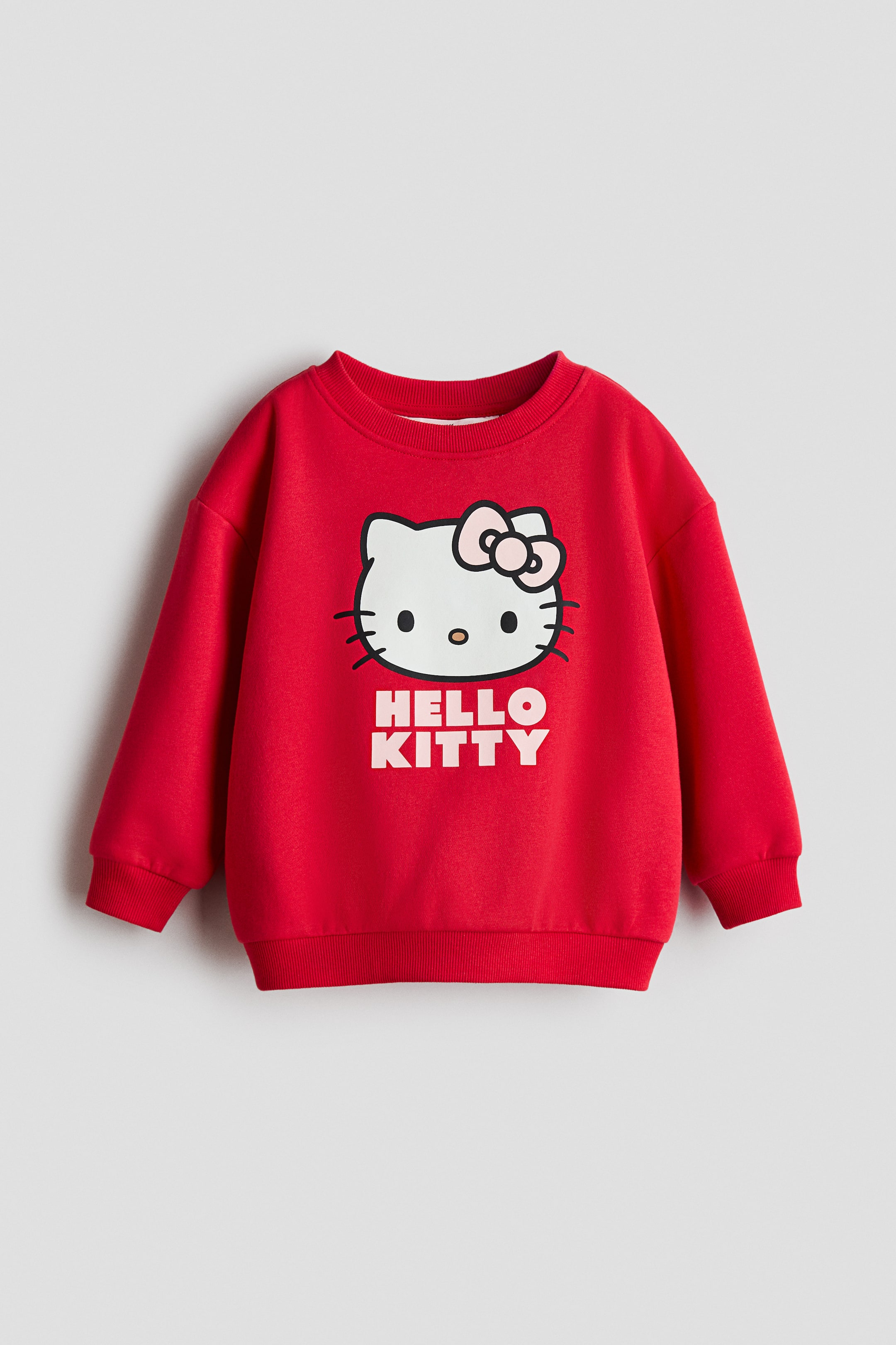 View larger image: A red jumper showcases a large white Hello Kitty graphic on its front, featuring her head adorned with a pink bow and the text "HELLO KITTY" beneath. The jumper is crafted with a ribbed crew neck, ribbed cuffs, and a ribbed hem.