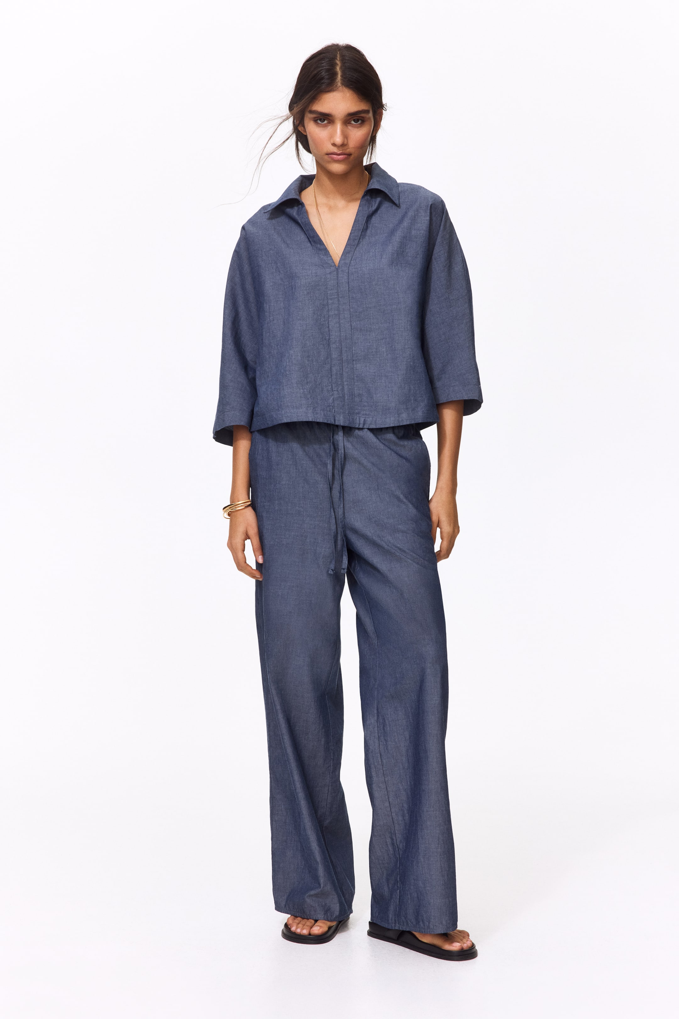 View larger image: The model is wearing loose-fit trousers crafted from a cotton weave. These trousers boast a high waist with covered elastication, a tied drawstring, diagonal side pockets, and straight legs.