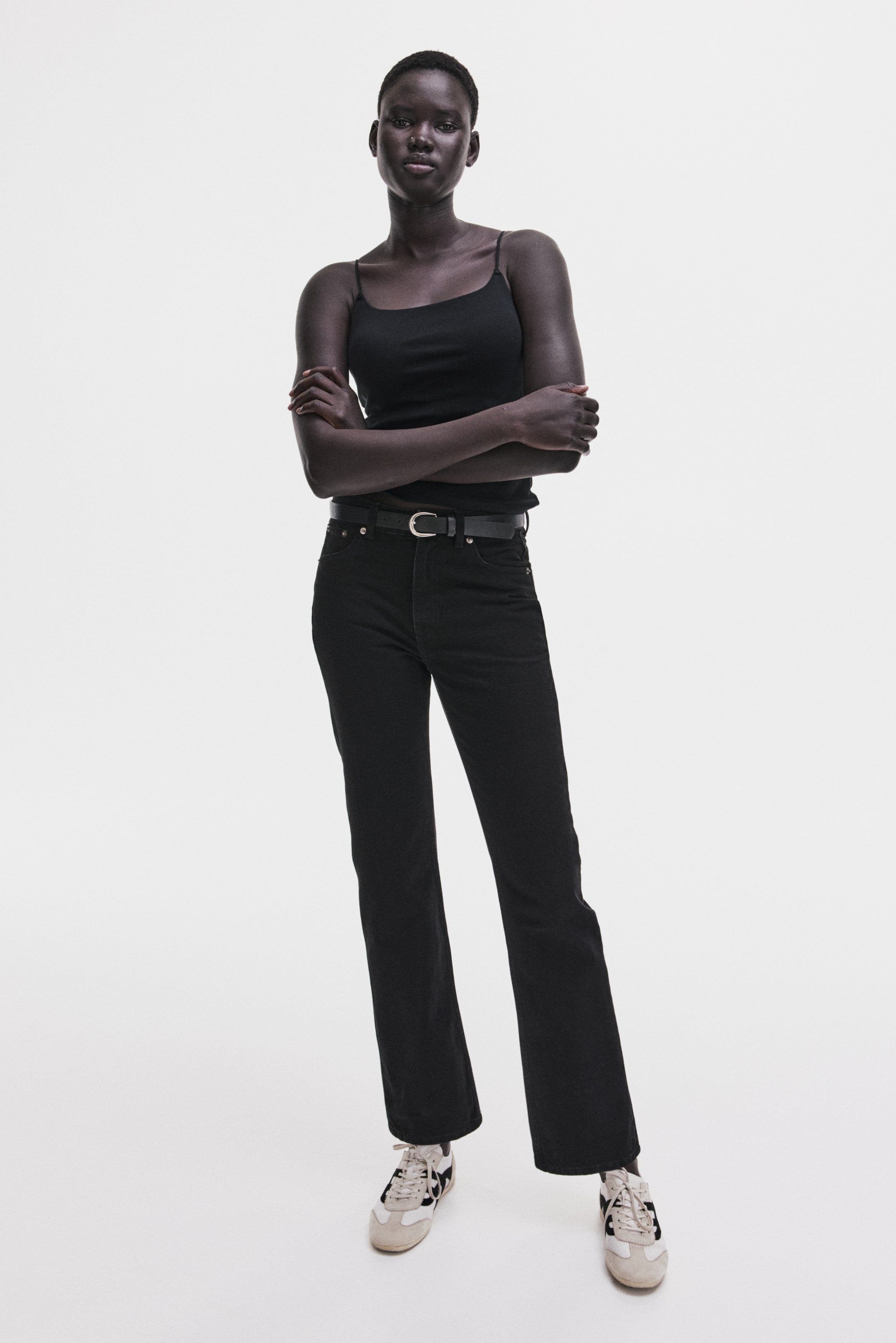 View larger image: A black spaghetti-strap tank top appears fitted and is tucked into high-waisted black jeans. The straight-leg jeans feature a black belt with a silver-toned buckle, while light-colored sneakers with black accents complete the outfit.
