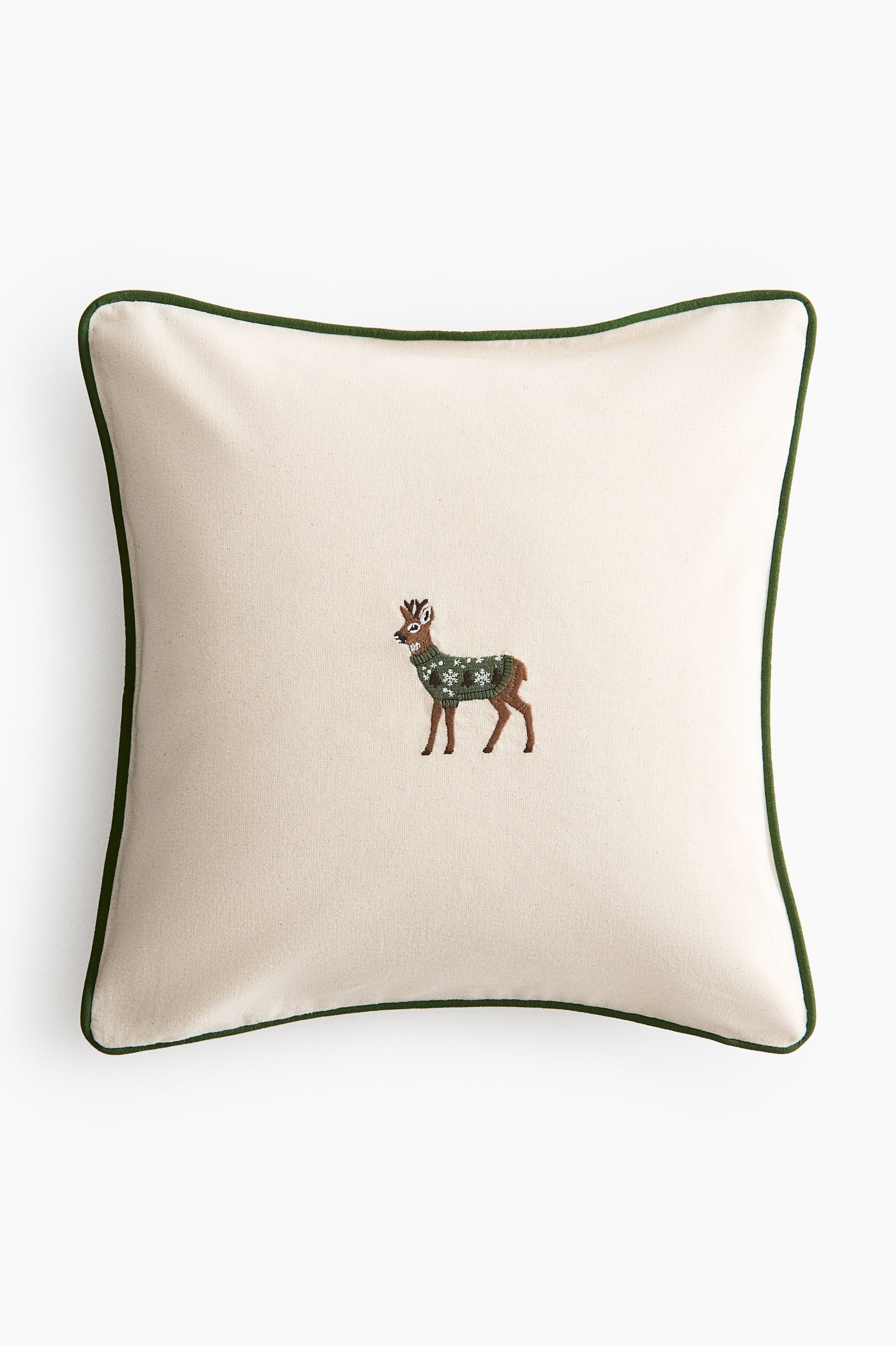 Agrandir l'image: Un coussin carré est recouvert d’un tissu blanc cassé avec un passepoil vert foncé sur ses bords. Un cerf brodé est placé au centre, tourné vers la gauche, portant un pull vert orné de motifs de flocons de neige blancs.