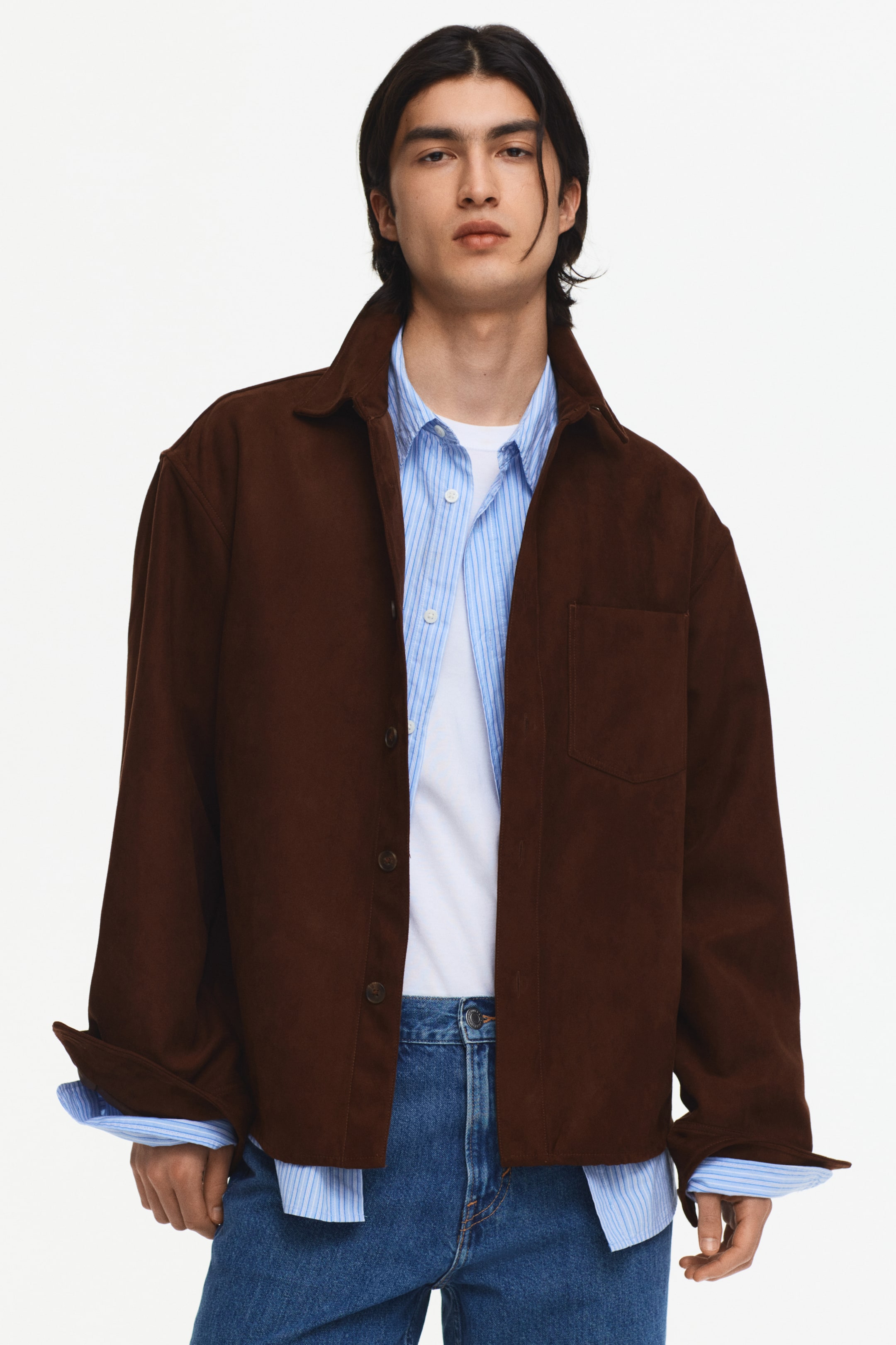 Se større billede: En brun overshirt i ruskind-look præsenteres fra en frontvendt vinkel, med en spids krave, en knaplukning med mørke knapper og en enkelt brystlomme på venstre side. De lange ærmer er rullet op, hvilket afslører en lyseblå stribet skjorte med krave, som bæres under og lagvis over en hvid t-shirt.