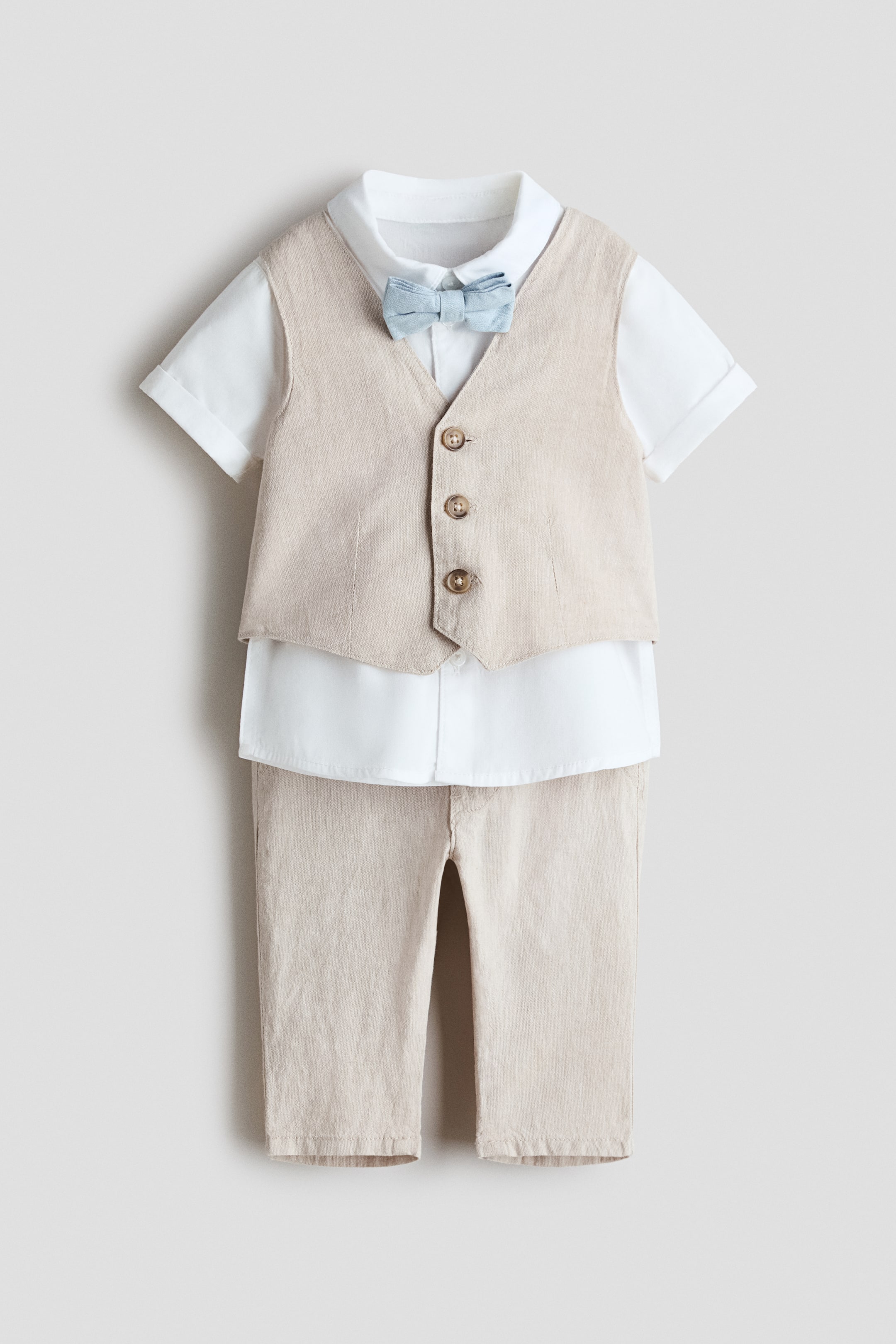 Ver imagen más grande: Se muestra el conjunto de vestir para niño VISTA FRONTAL, con un chaleco beige claro con tres botones marrones en la parte delantera y pantalones a juego. El chaleco está superpuesto a una camisa blanca de manga corta con cuello de solapa y vueltas cosidas en los puños, con una pajarita azul claro preanudada que acentúa el cuello.