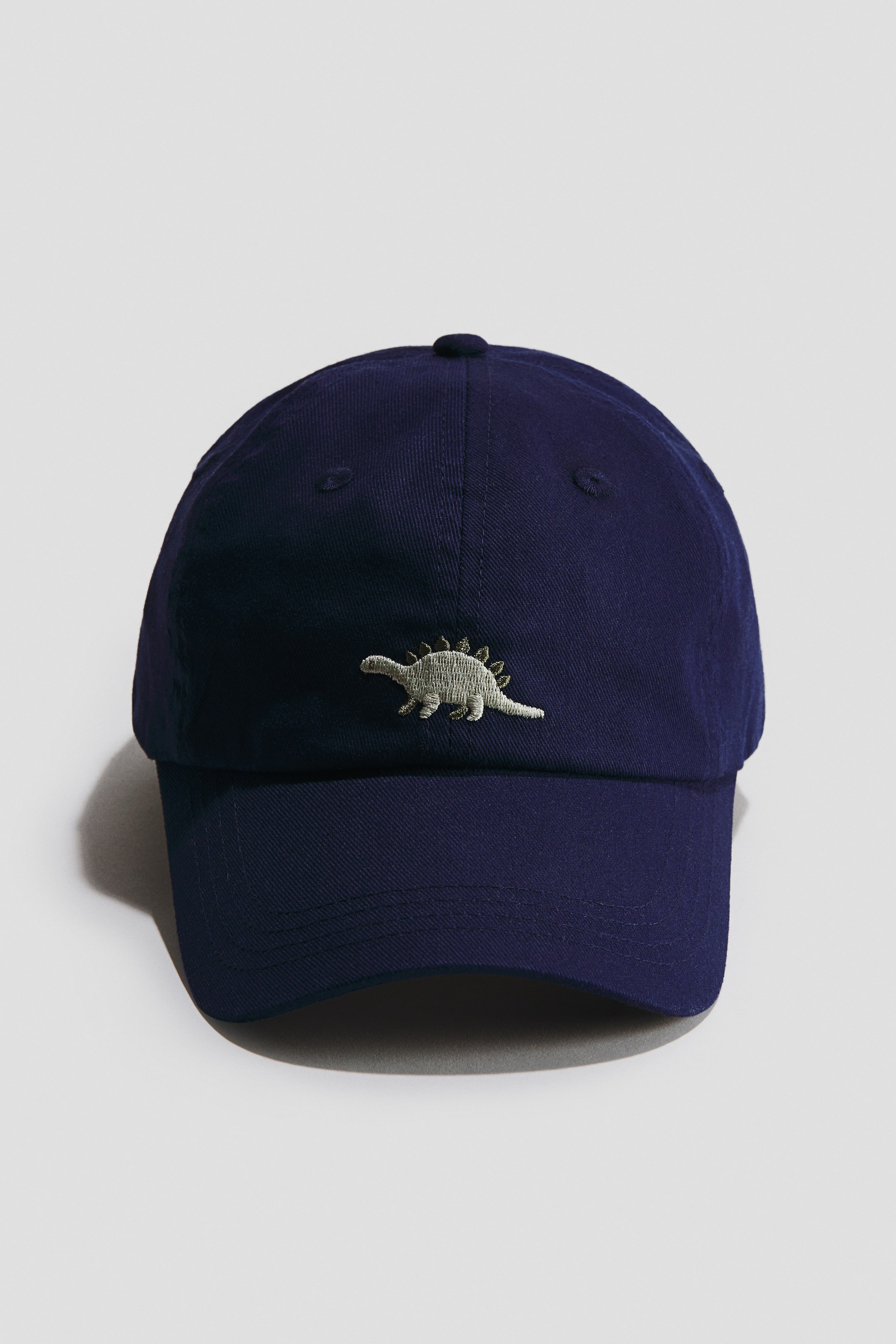 Visualizza immagine più grande: Un cappello in twill di cotone blu navy presenta un motivo ricamato di stegosauro in grigio chiaro sul davanti, con occhielli ricamati visibili sulla corona.
