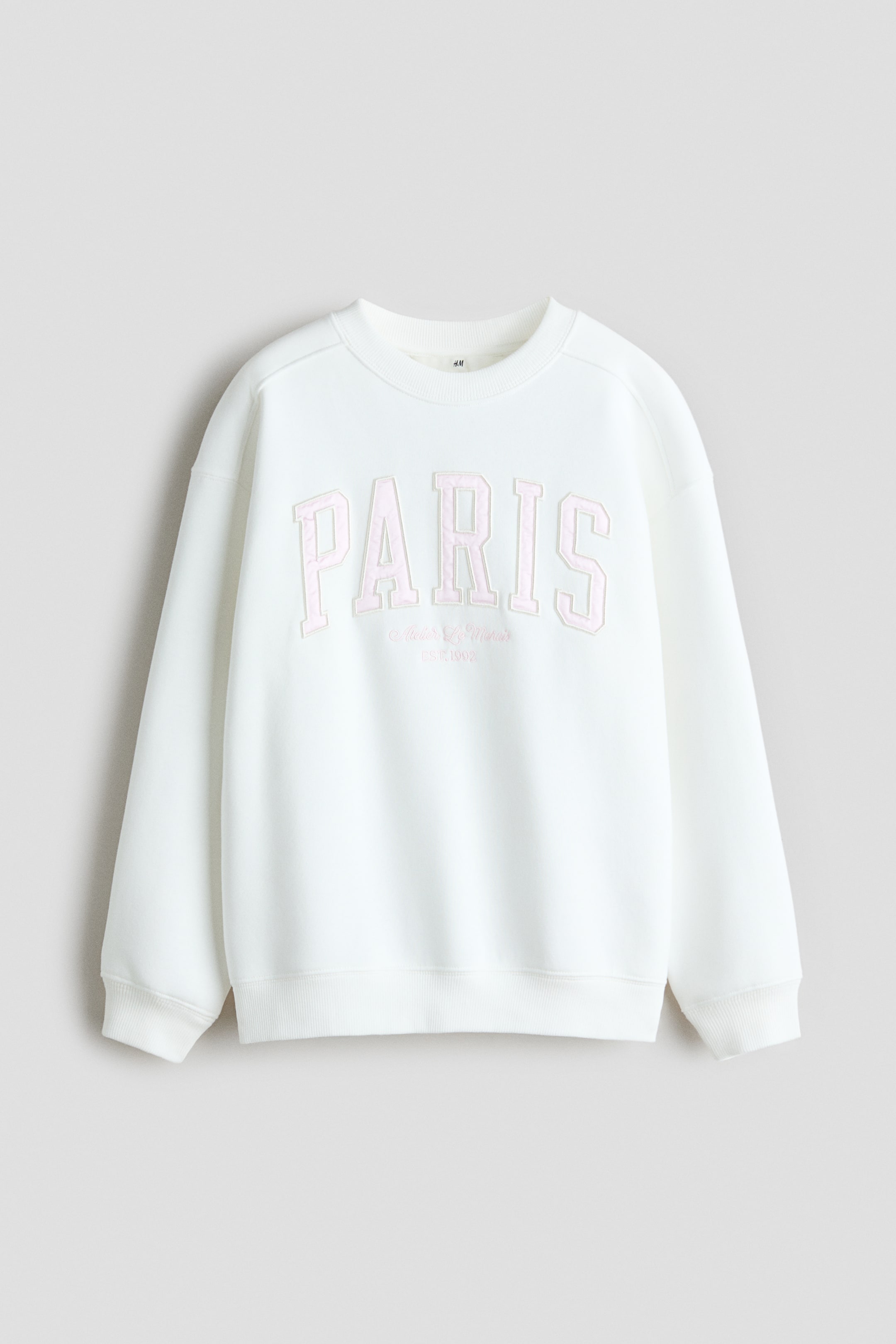 View larger image: A loose-fitting, off-white sweatshirt is shown from the front, featuring a round, rib-trimmed neckline, dropped shoulders, and long sleeves with ribbed cuffs and a ribbed hem. The front displays a collegiate-style graphic design with the word "PARIS" printed in light pink block letters.