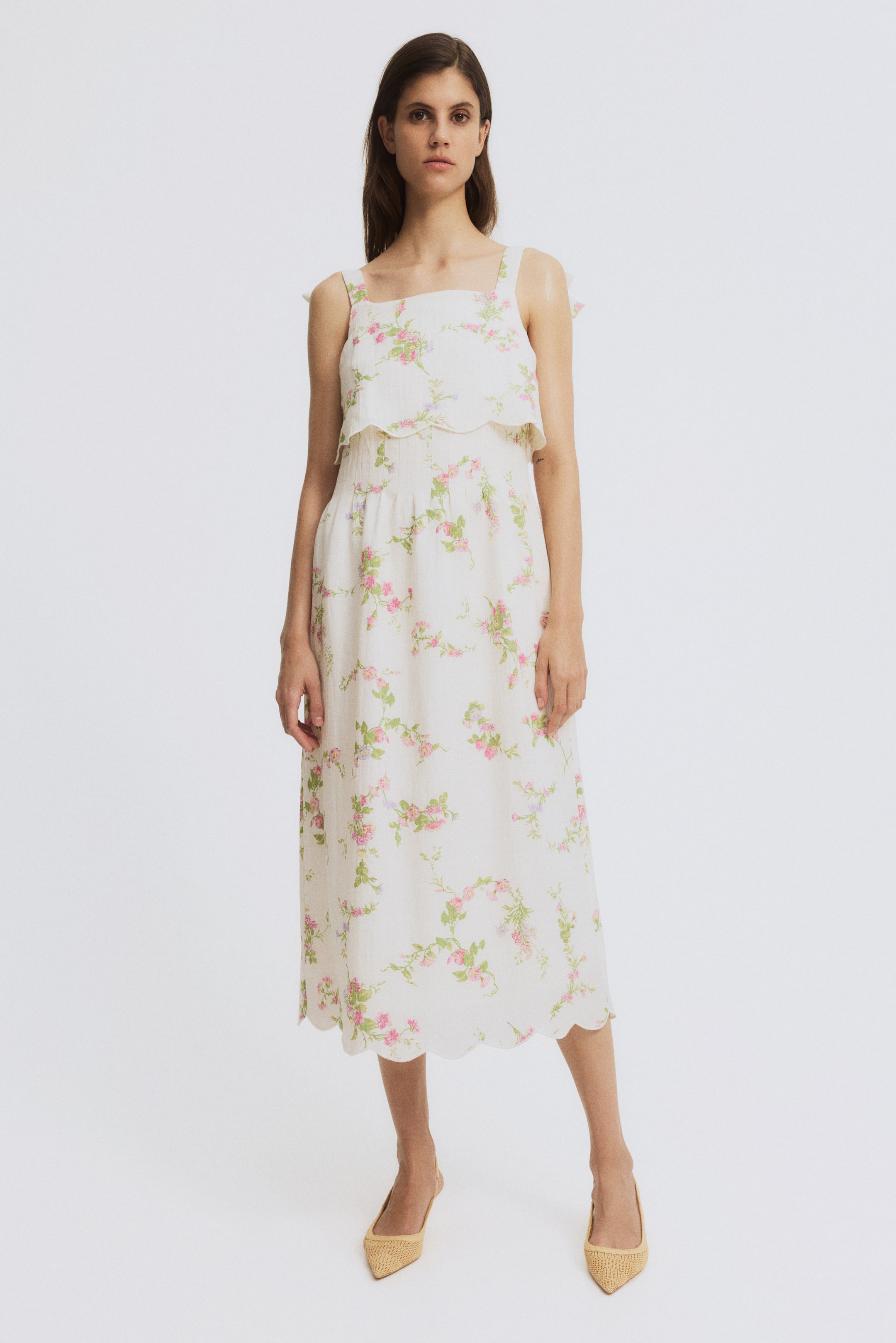 View larger image: A cream-colored midi dress features a pink and green floral print, a square neckline with wide shoulder straps, and a scalloped flounce overlay at the bust, flowing into a gathered skirt with a scalloped midi hemline.