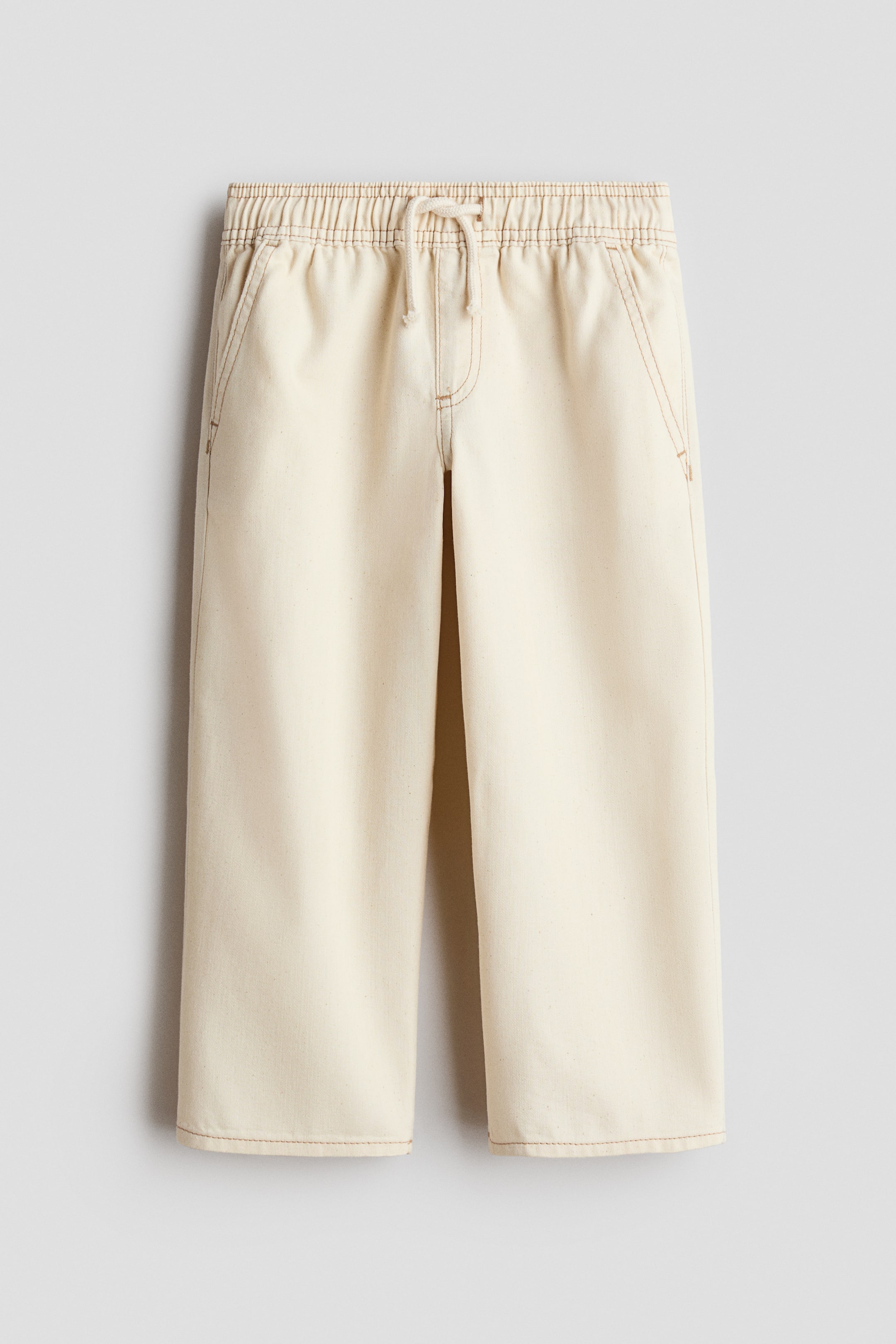View larger image: Cream-coloured denim joggers are hanging in a front view, displaying a loose, straight-leg silhouette with contrasting tan stitching. They feature an elasticated waistband with a drawstring, a mock fly, and diagonal side pockets.