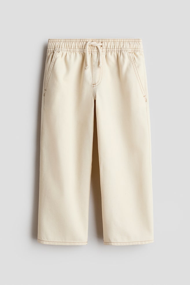 Cream-colored denim joggers are hanging in a front view, displaying a loose, straight-leg silhouette with contrasting tan stitching. They are showcasing an elasticated waistband with a drawstring, a mock fly, and diagonal side pockets. - 1