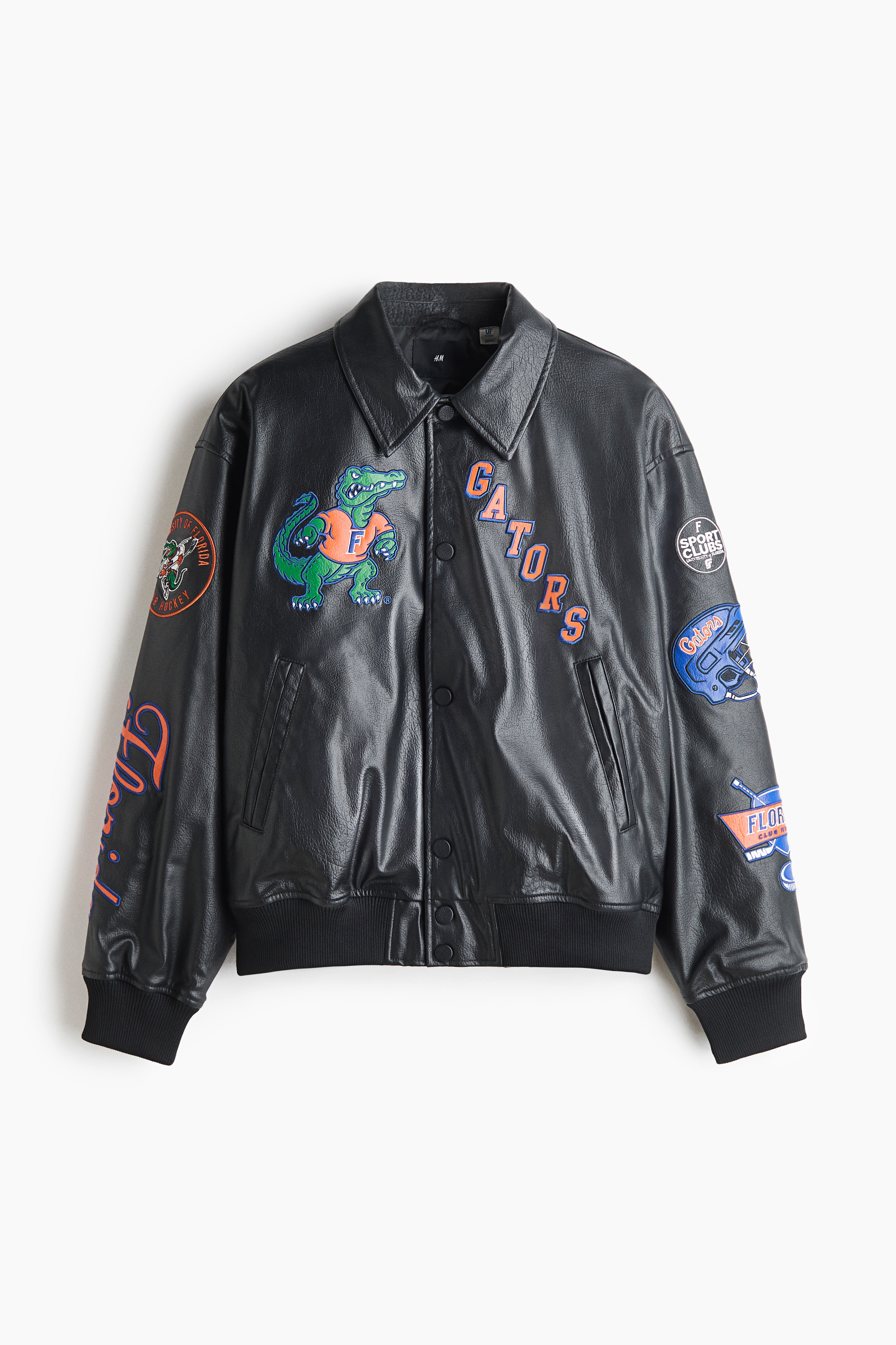 Une veste de baseball noire en tissu enduit, ornée d’un alligator brodé en guise de mascotte et du lettrage « GATORS », est portée par le mannequin. La veste présente un col, une fermeture par boutons-pression sur le devant et des bordures côtelées, et elle est superposée à une chemise en denim foncé et à un t-shirt gris.