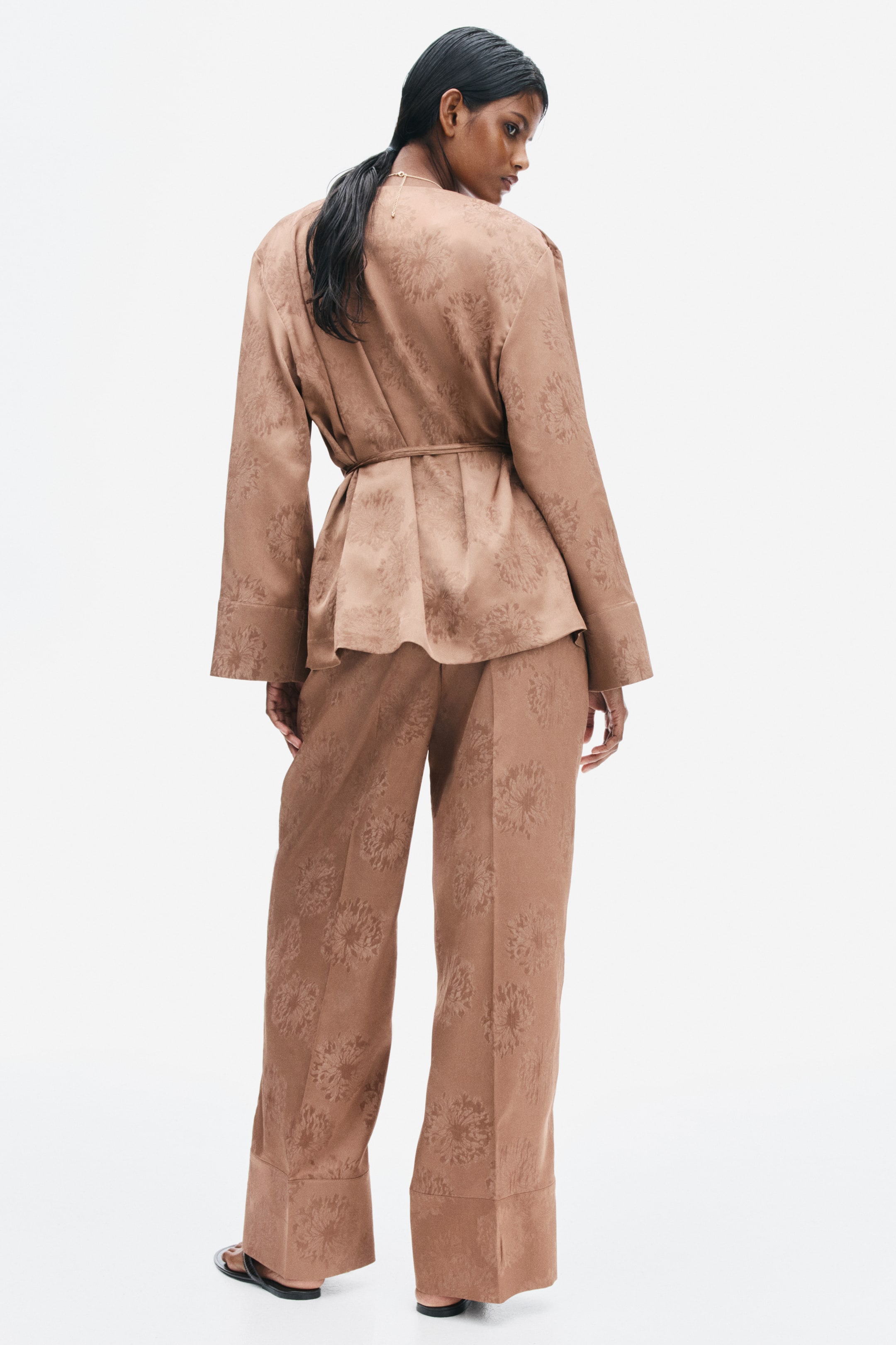 View larger image: Loose-fit, wide-leg trousers in bronze jacquard-weave satin are worn by the model, featuring pressed creases down the back of the legs. The trousers are paired with a matching long-sleeved wrap jacket in the same fabric.