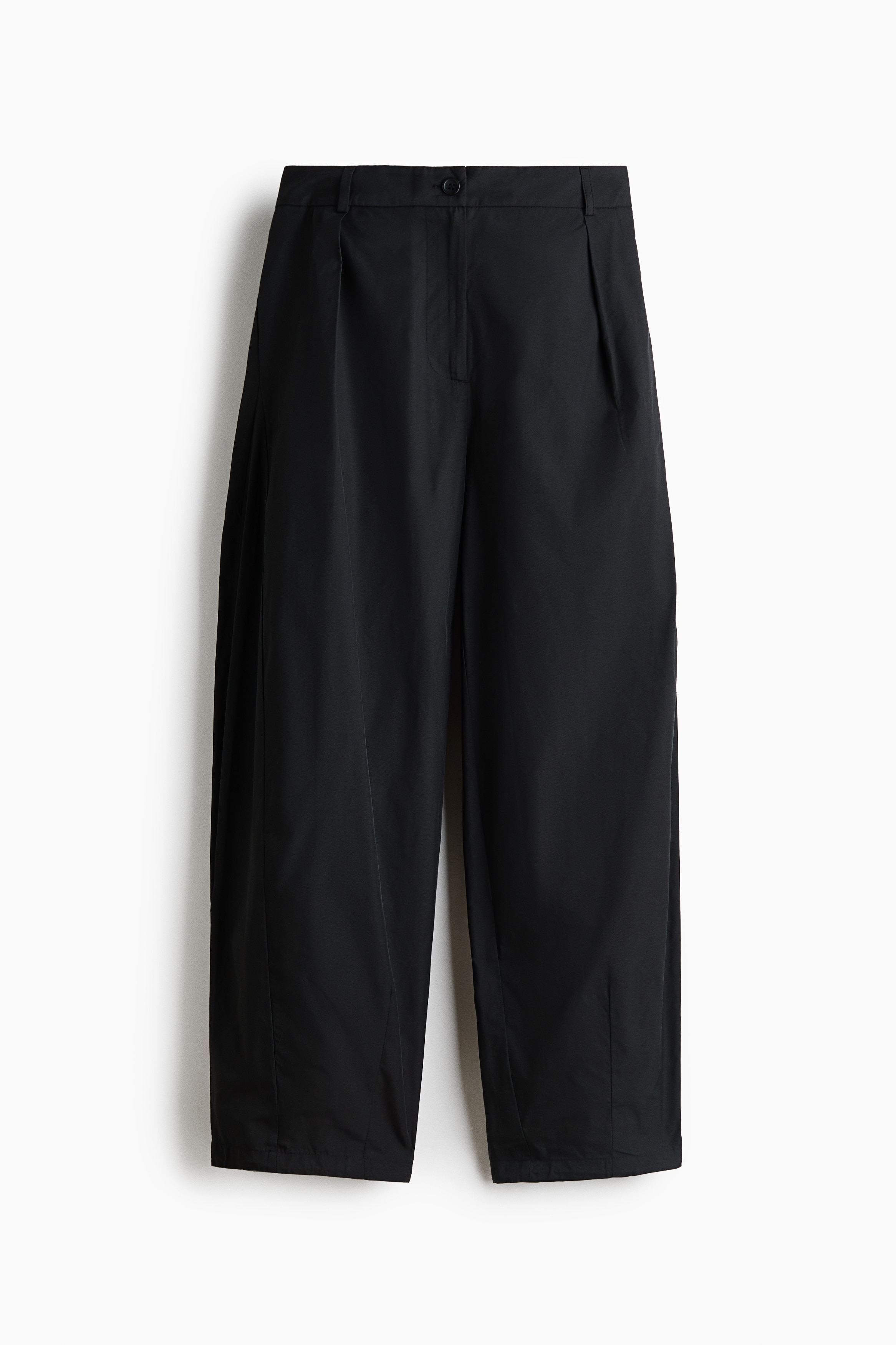 Pantaloni neri dal taglio loose fit in twill, con chiusura a bottone, pieghe nella parte superiore e gamba barrel, indossati dalla modella. I pantaloni sono abbinati a un top bianco a maniche lunghe infilato nei pantaloni e a un maglione nero drappeggiato sulle spalle.