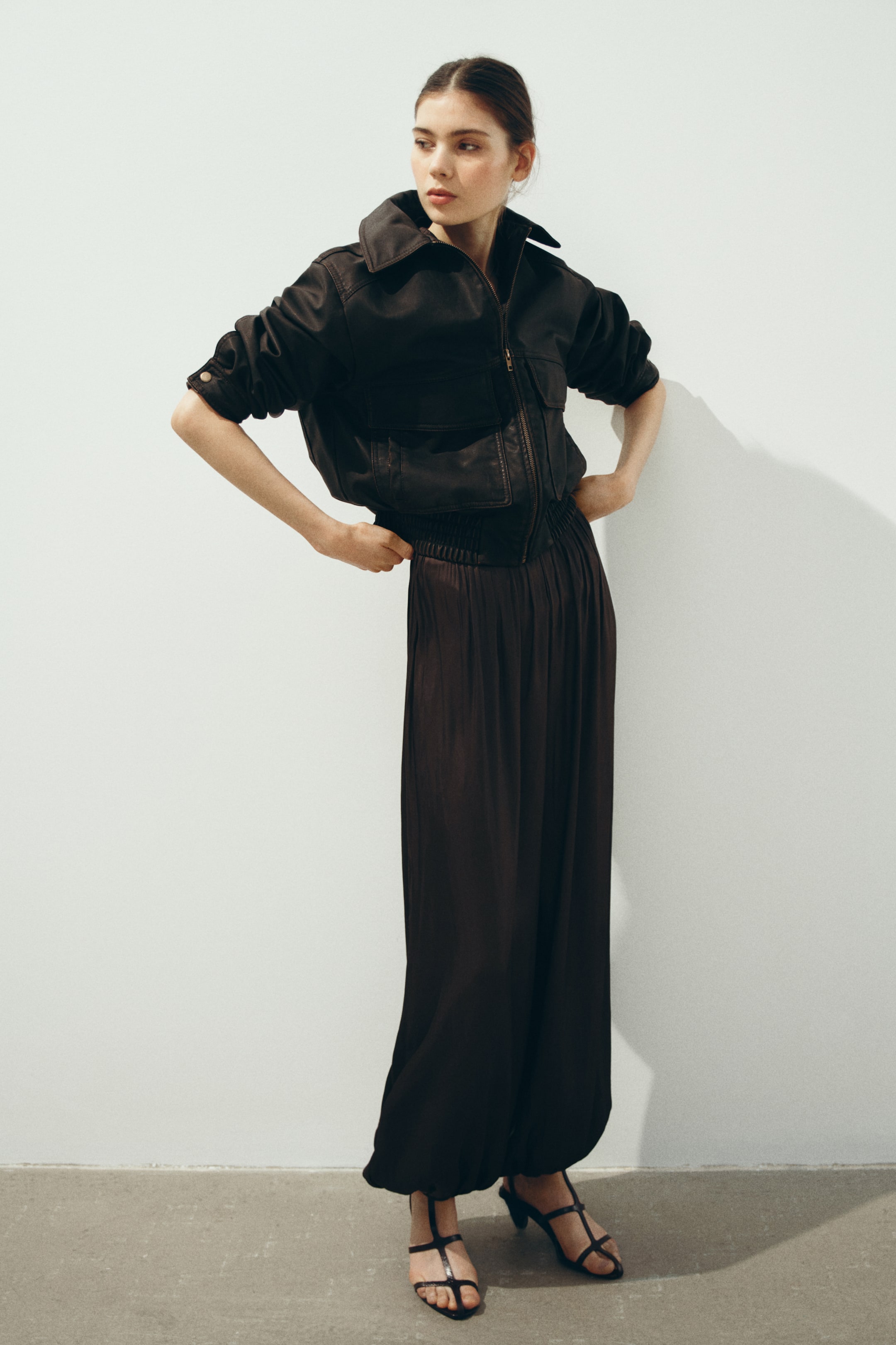 View larger image: A dark, cropped utility jacket is worn, featuring a wide collar, a front zipper, two chest flap pockets, and a shirred hem. The sleeves are rolled up, revealing buttoned cuffs, styled with dark, flowing wide-leg pants showcasing a shirred waistband and gathering at the ankles, along with dark strappy sandals.