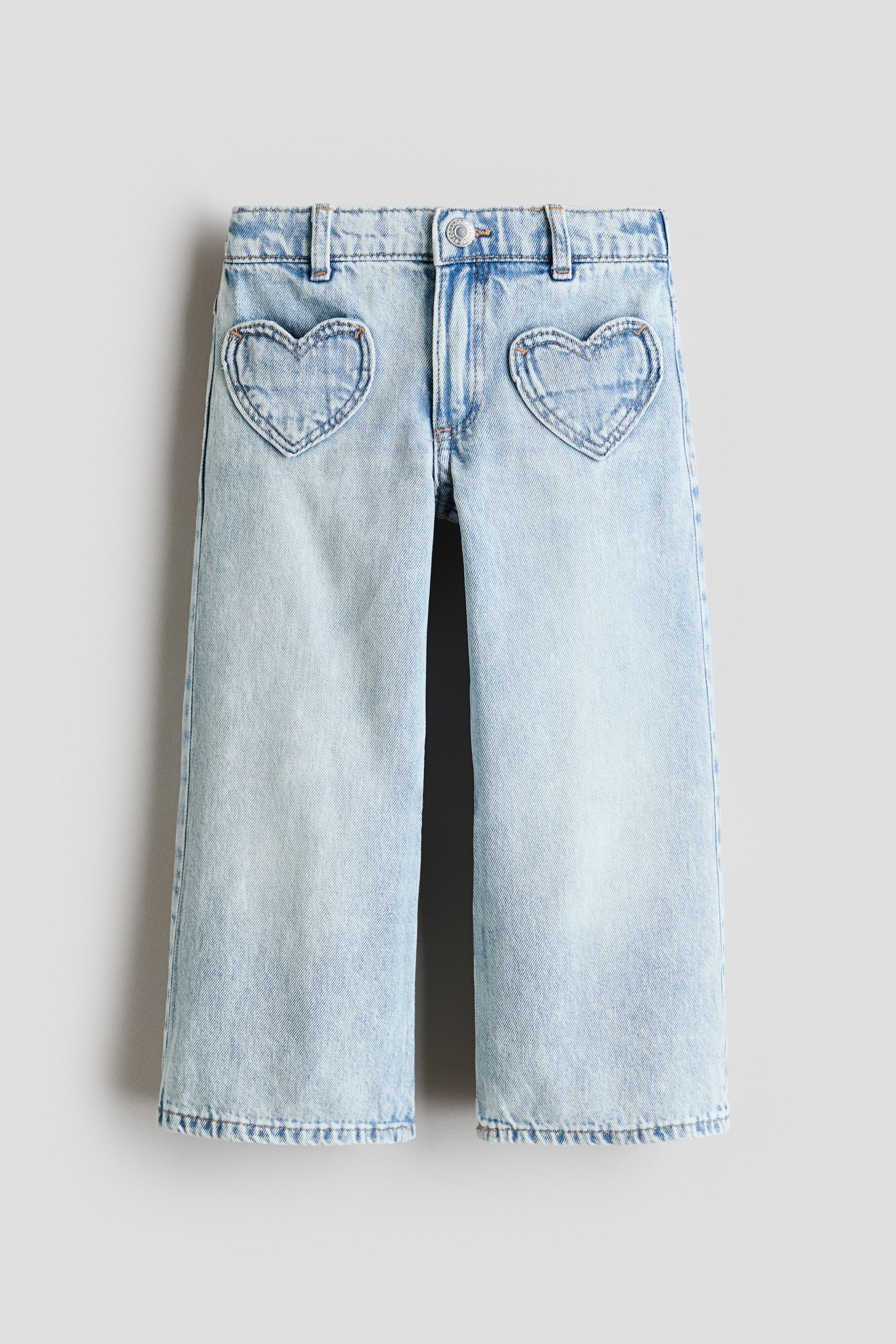 View larger image: Light blue denim jeans showcase a wide-leg cut and two heart-shaped front pockets, outlined with visible stitching. These jeans feature a light wash with subtle distressing and a visible button closure at the waistband.