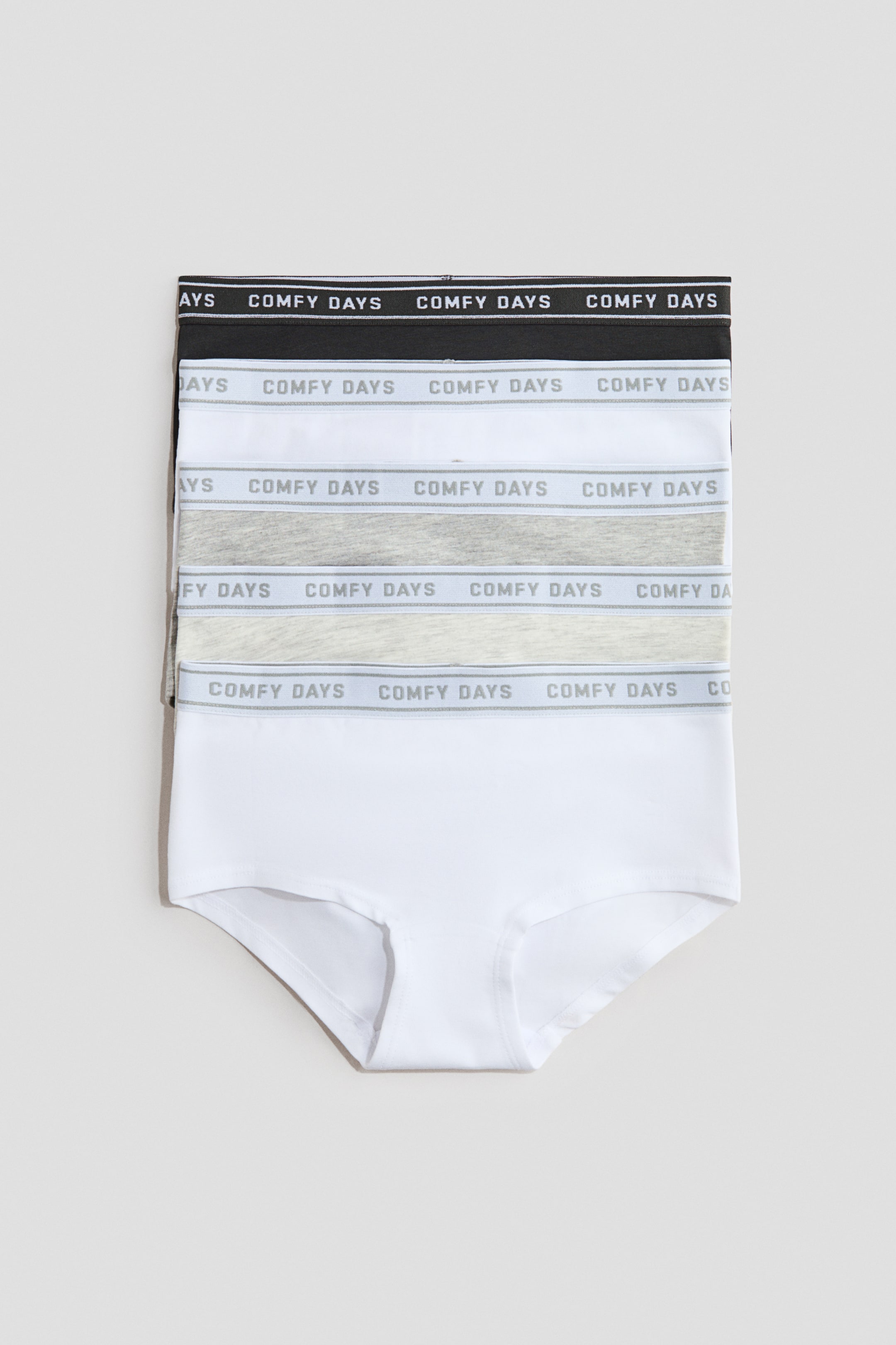 Visualizza immagine più grande: Una pila di cinque paia di biancheria intima presenta la vista frontale, con fasce elastiche in vita recanti la scritta "COMFY DAYS". La pila mostra paia nei colori nero, bianco, grigio chiaro e grigio scuro, con l'ultimo paio completamente visibile di colore bianco.