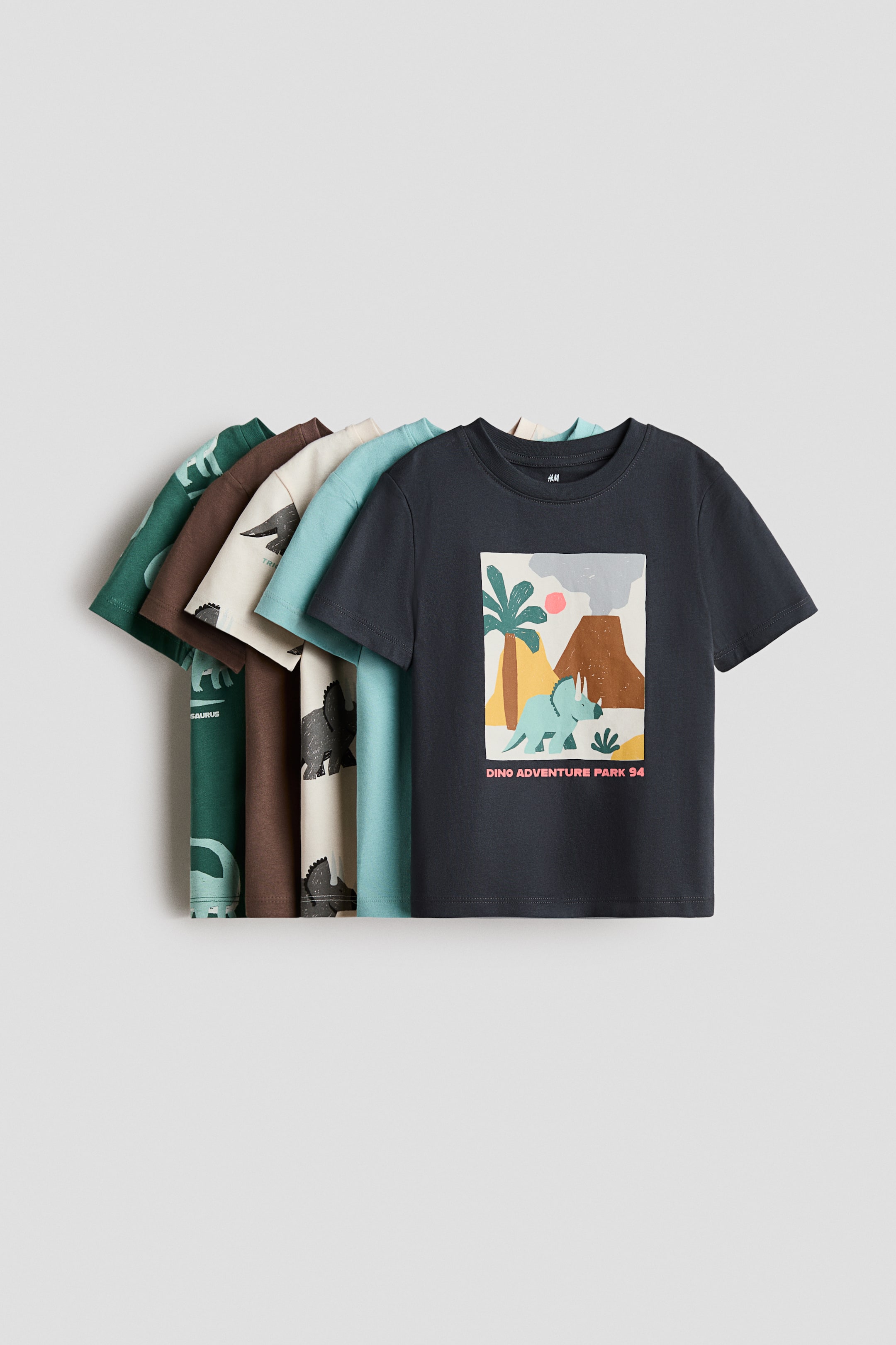 View larger image: Five classic T-shirts are showcased, stacked with the foremost dark gray T-shirt facing forward, featuring a rectangular print motif on the front. This design depicts a dinosaur, a volcano, a palm tree, and the text "Dino Adventure Park '94." The partially visible T-shirts behind also display various print motifs, and all visible T-shirts feature round, ribbed necklines.