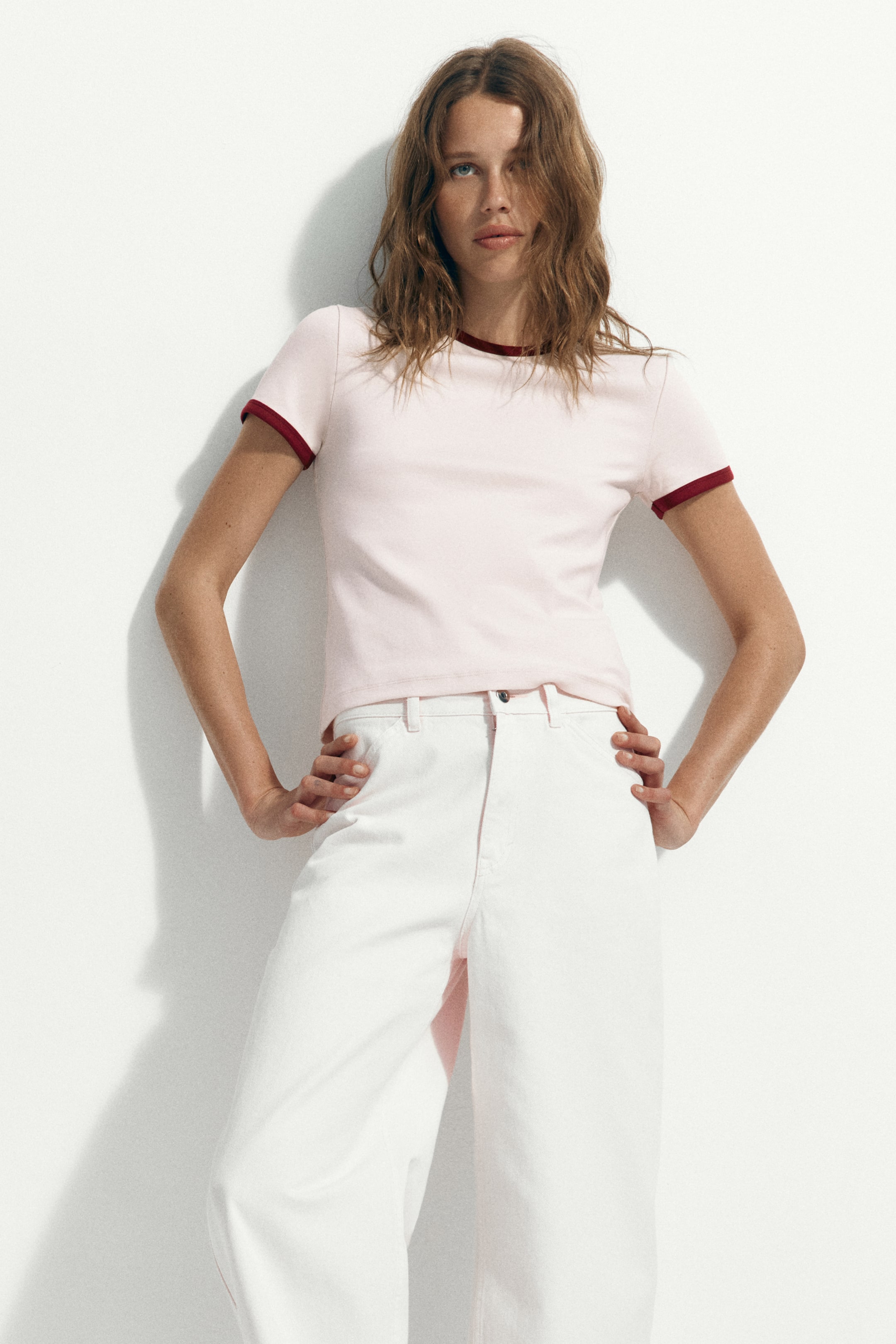Voir une image plus grande: Un t-shirt ringer rose pâle présente une bordure rouge foncé le long de son col rond et de ses poignets de manches courtes, s'ajustant près du corps et orienté vers l'avant pour mettre en valeur son design.