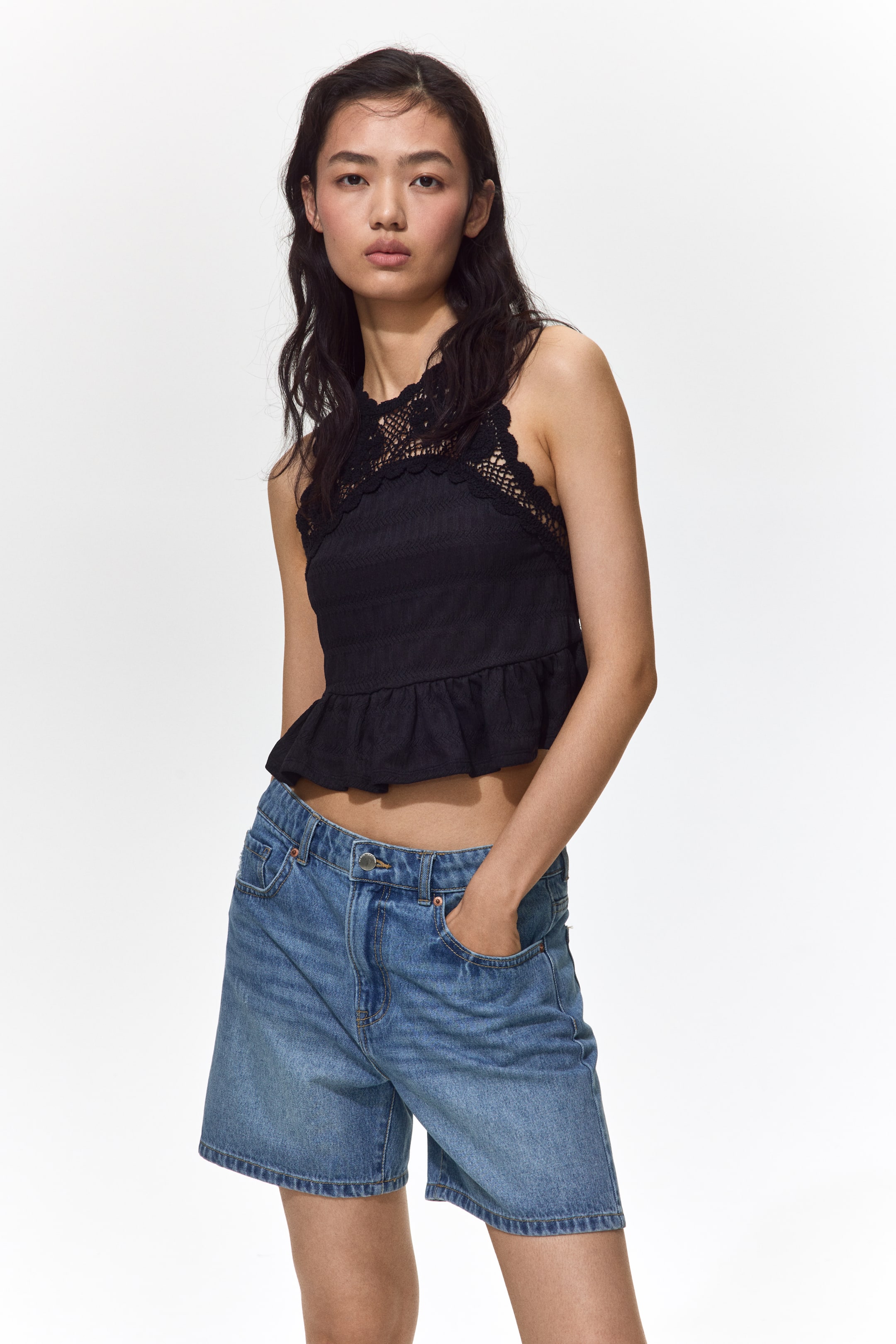 Grotere afbeelding bekijken: Een zwarte mouwloze top met hoge hals, voorzien van ingewikkeld haakwerk kant over de borst en schouders, een getextureerde body en een gerimpelde peplumzoom. Het wordt gedragen met lichtgewassen denim Bermuda shorts, die een hoge taille en een ontspannen pasvorm tot halverwege de dij laten zien.