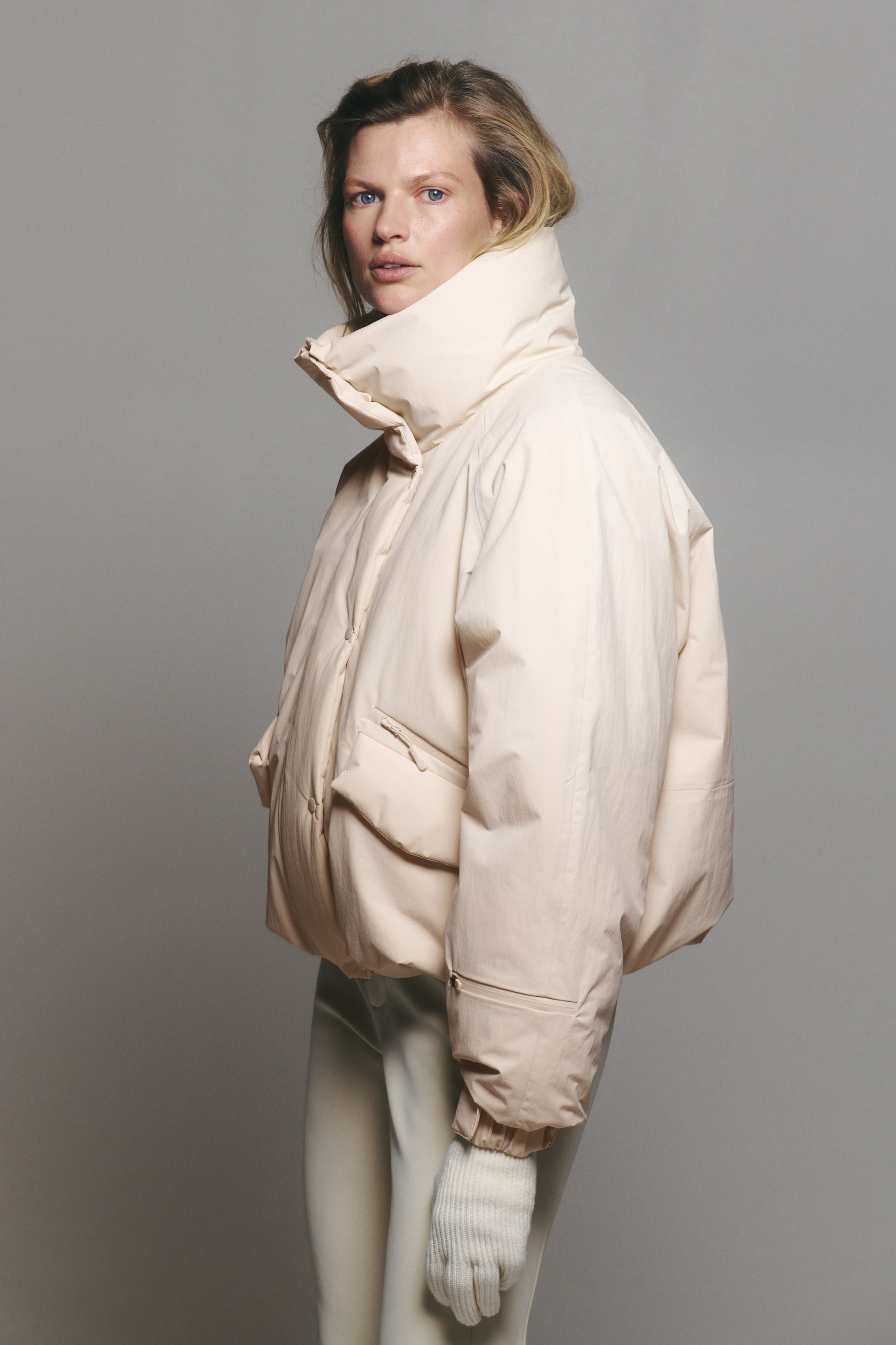 View larger image: A cream-coloured, oversized puffer jacket featuring a high stand-up collar, snap-button front, and zippered flap pockets is being worn by the model. The jacket is styled with matching off-white trousers and a white ribbed-knit glove.