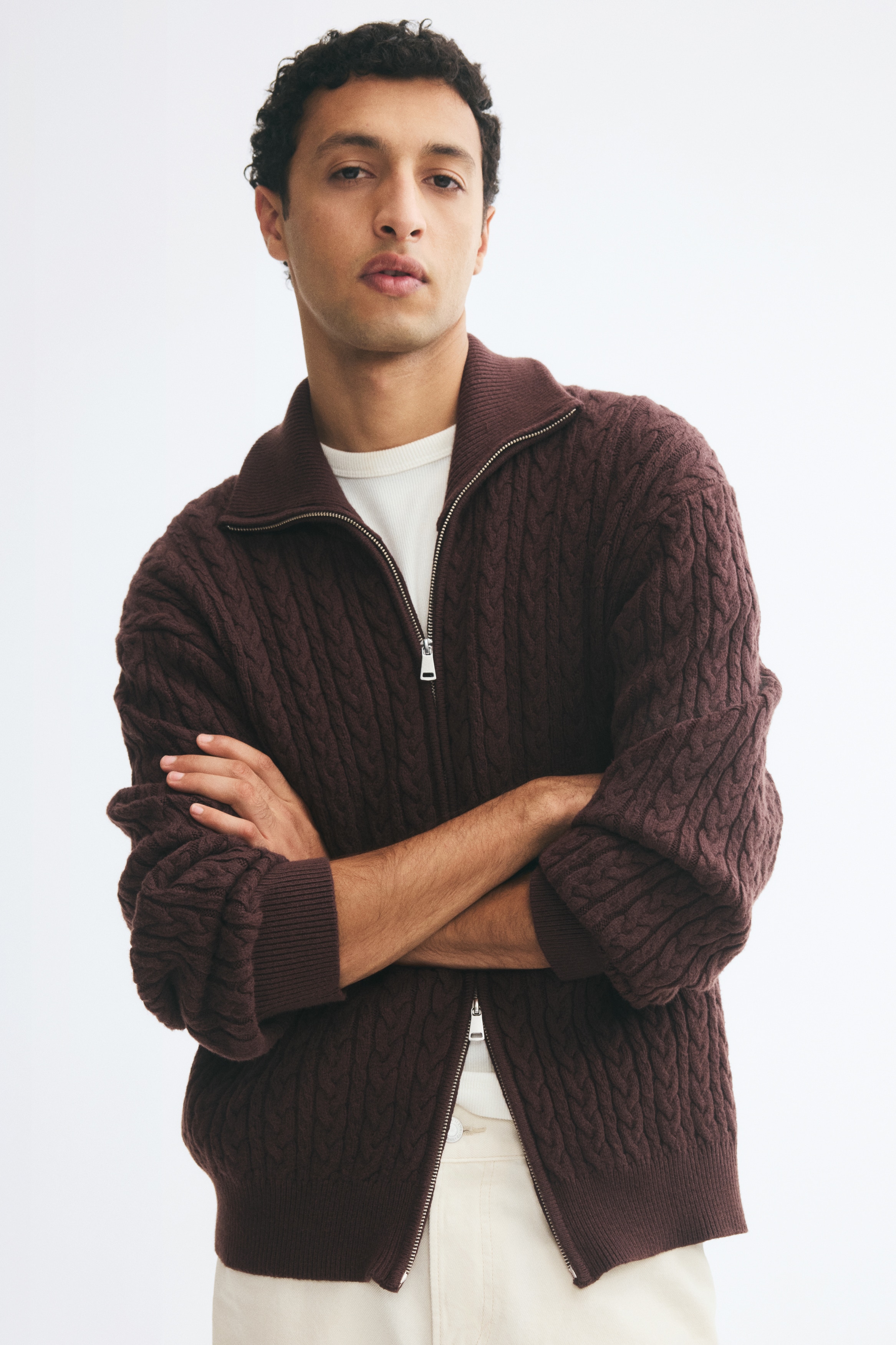 A dark brown cable-knit cardigan is facing front, featuring a high ribbed collar and long sleeves. It is displaying a silver-toned two-way front zipper, ribbed cuffs, and a matching ribbed hem.