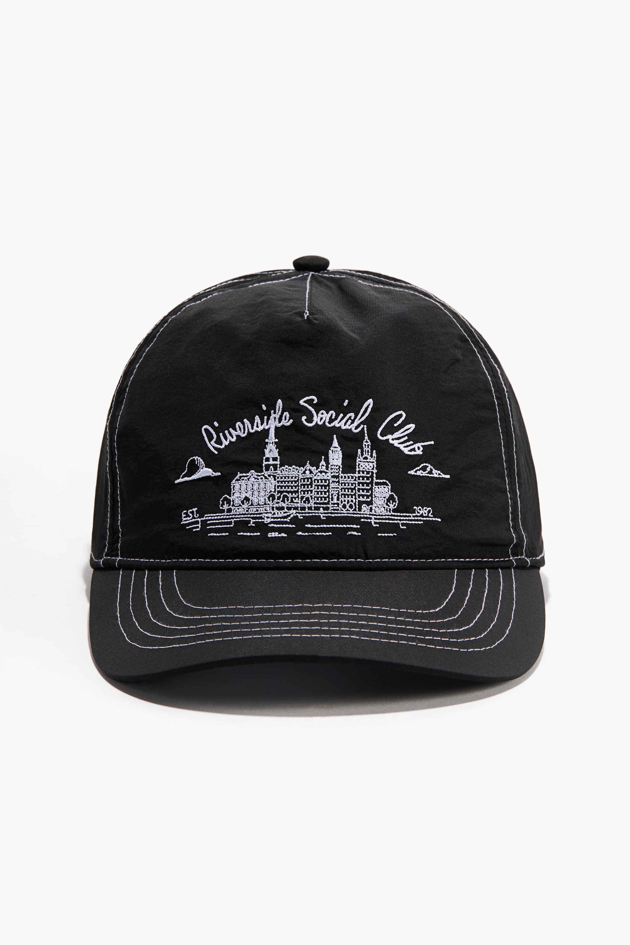 拡大画像を表示: 黒い野球帽は、前面に「Riverside Social Club」の白い刺繍文字が、白い刺繍の都市風景デザインの上に表示されており、両側には「EST.」と「1982」が配置されています。パネルとフラットなつばには、白いコントラストステッチが施されています。