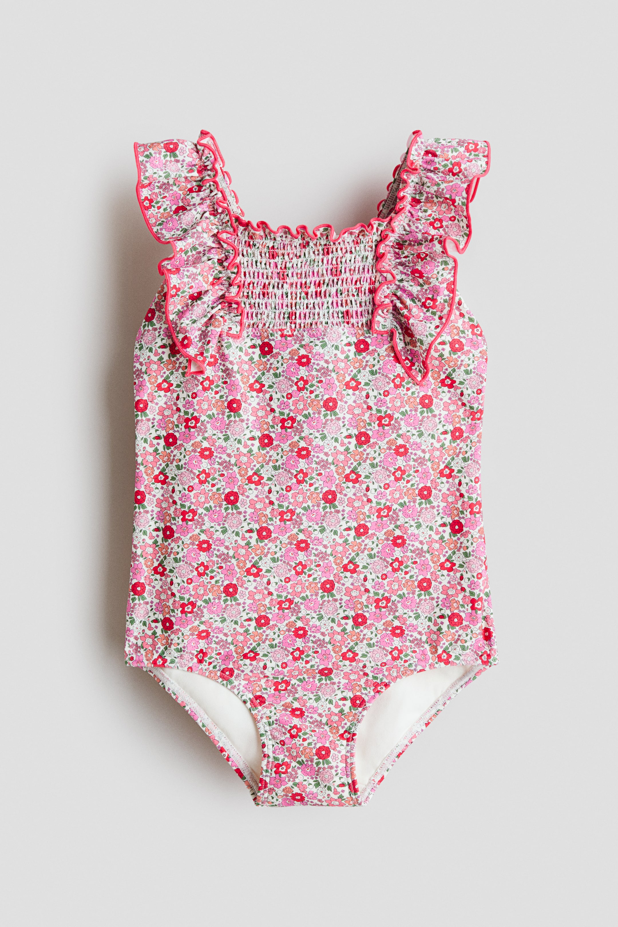 View larger image: The children's swimsuit is displayed from the front, showcasing a floral pattern of pink and red flowers accented with green leaves. It features a shirred bodice and ruffled shoulder straps, trimmed with vibrant pink piping.