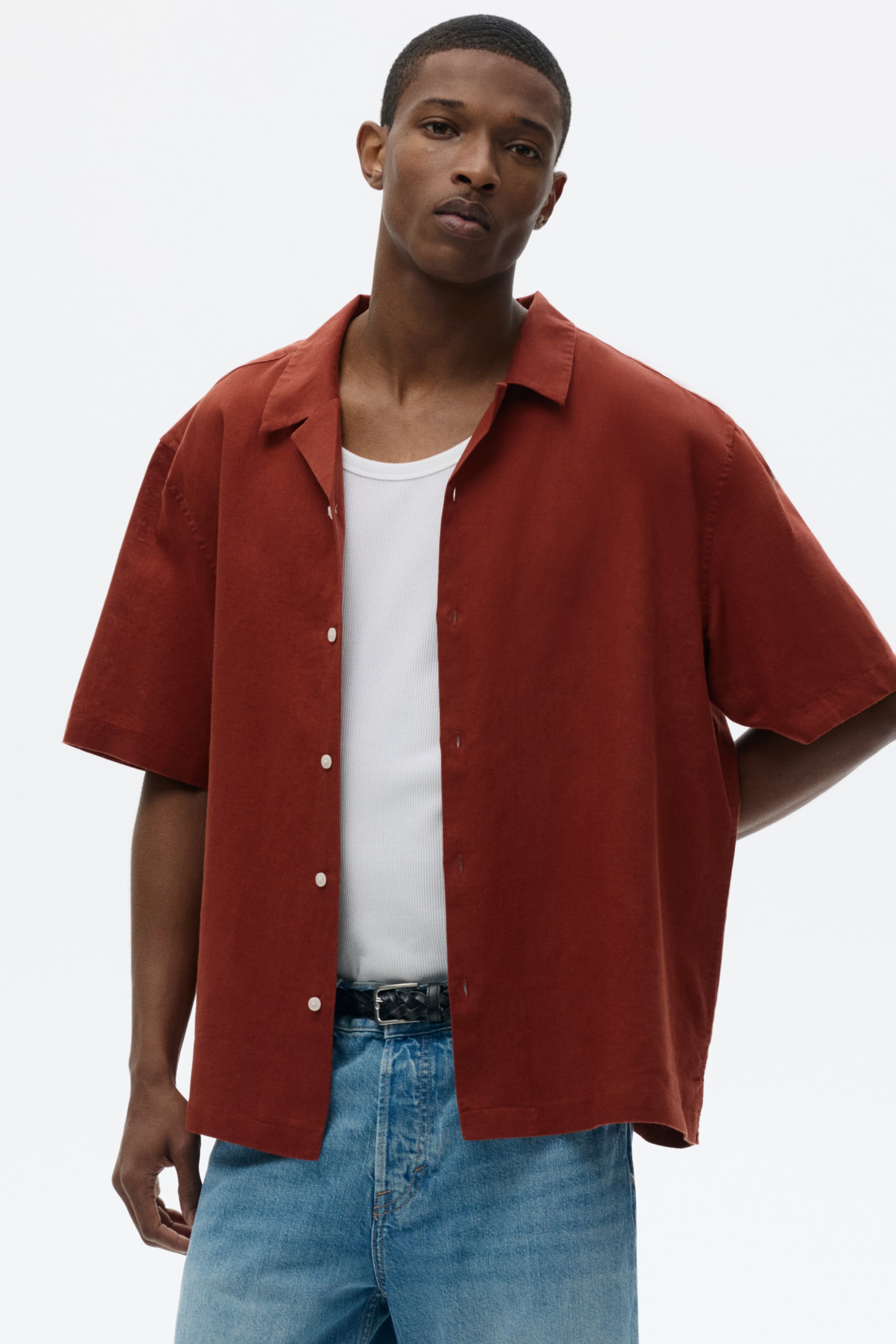 View larger image: A rust-colored short-sleeved shirt faces forward, featuring a spread collar and white buttons along the front placket. It is worn open over a white ribbed tank top, with light blue jeans and a black woven belt visible beneath the open front.