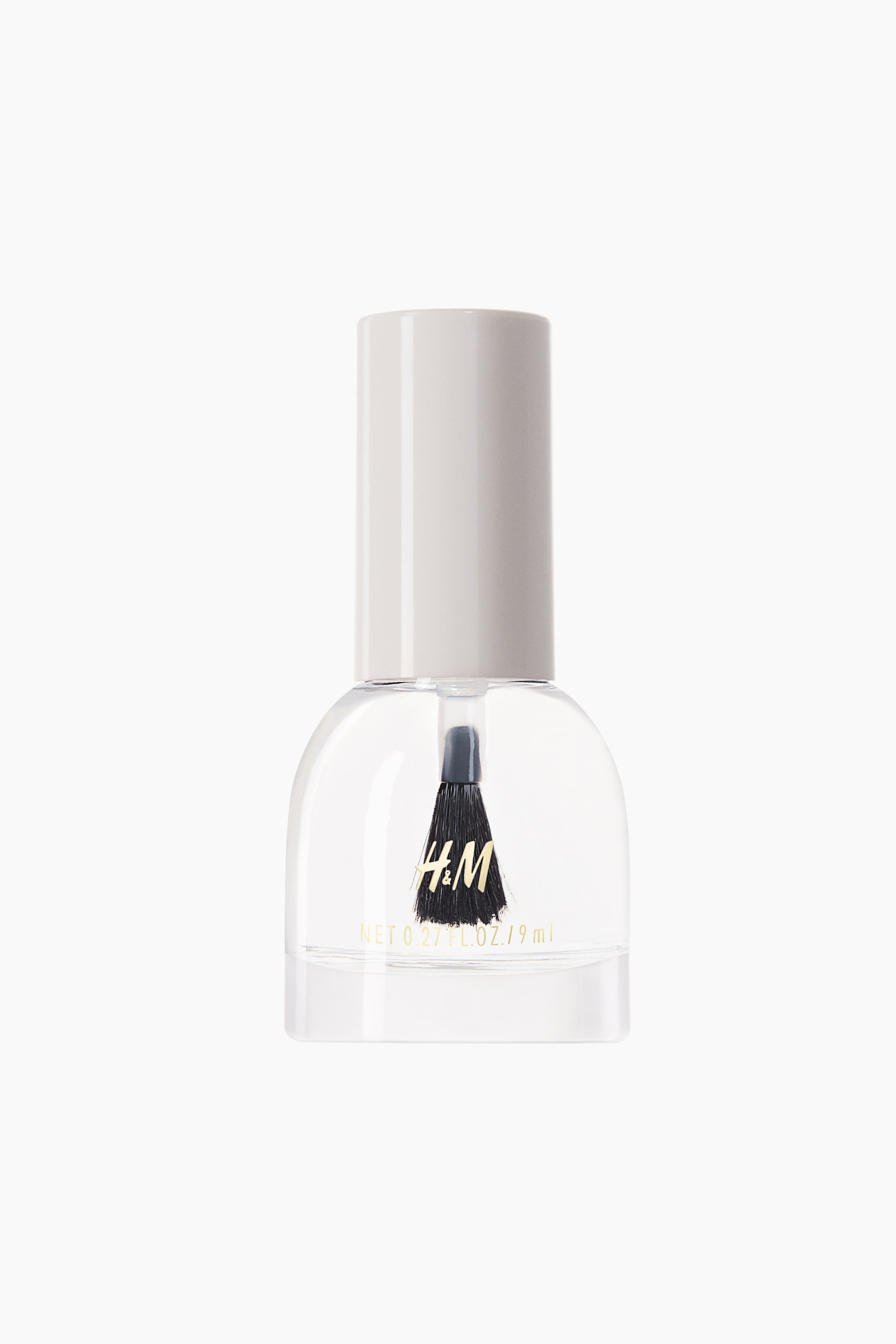 View larger image: A bottle of clear nail polish is standing upright, facing front, showcasing a rounded clear glass bottle filled with transparent liquid and topped with a white cylindrical cap. A black applicator brush is hanging inside the bottle, which is displaying gold branding text on the front.