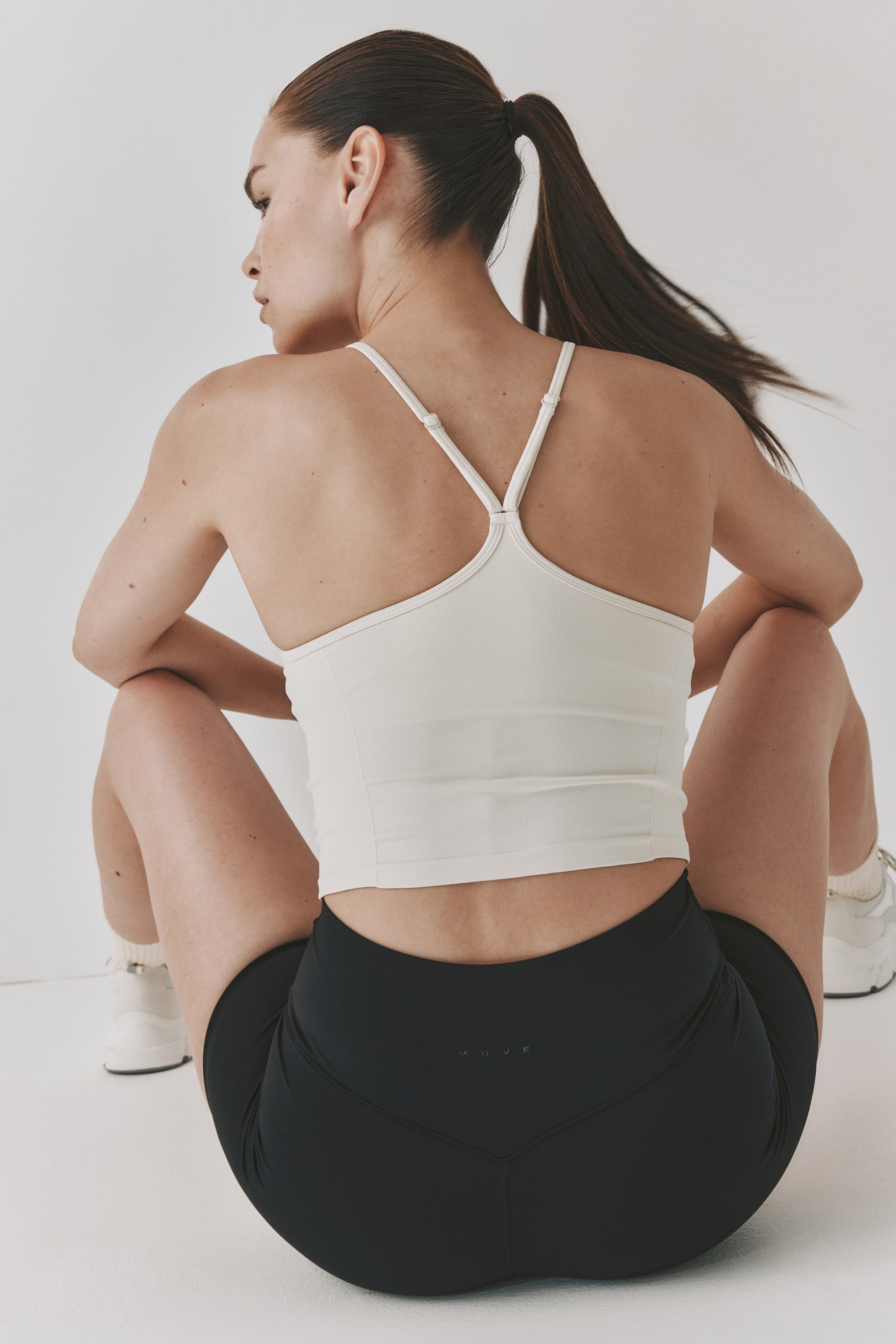 Grotere afbeelding bekijken: Een naadloze sporttop met DryMove? wordt gedragen door het model, met een lichtgekleurde, cropped silhouet en verstelbare spaghettibandjes die een Y-rug vormen. Het model zit met de rug naar de kijker toe, en draagt de top met zwarte sportshorts.
