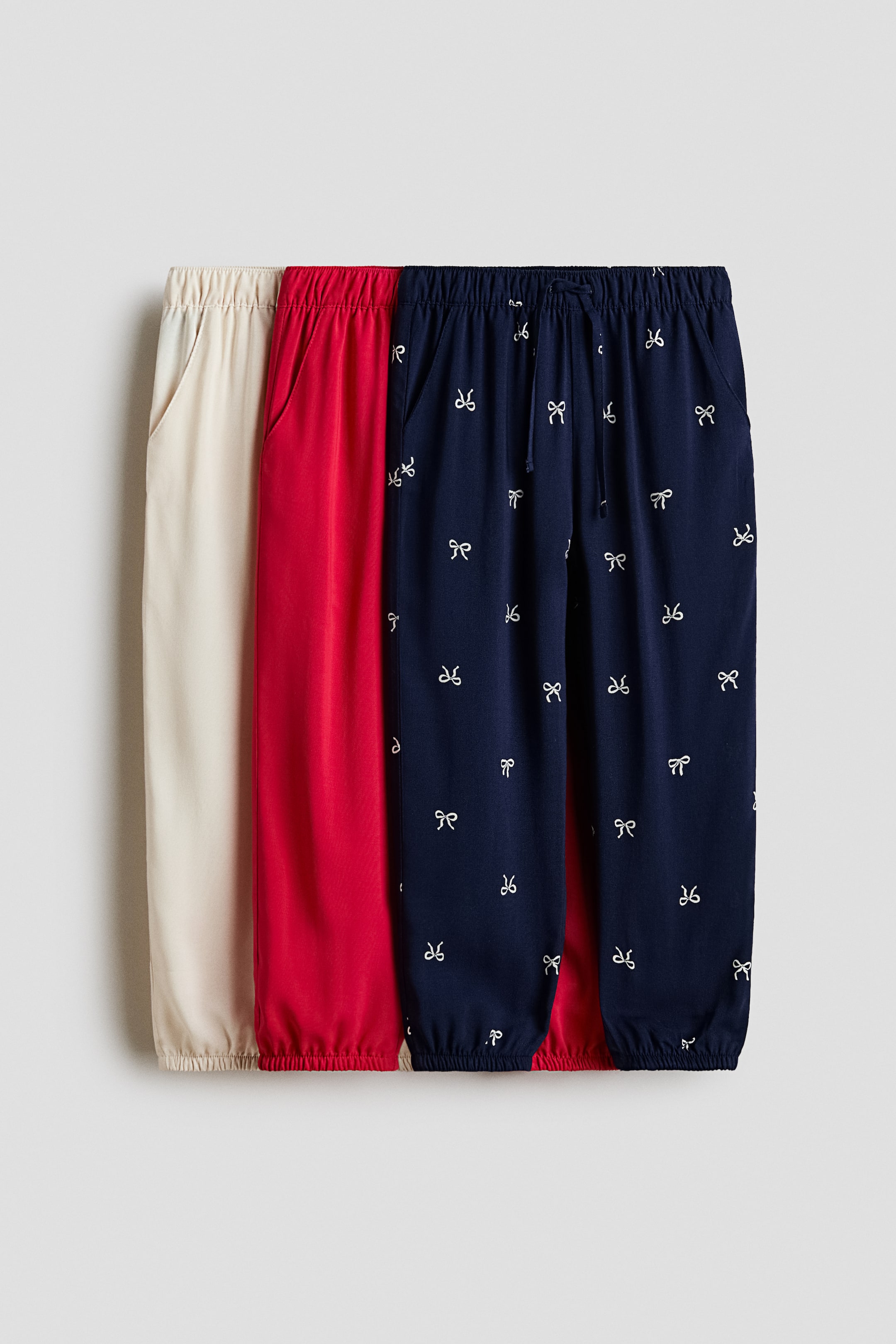 Agrandir l'image: Trois paires de joggeurs, confectionnées dans une toile de viscose légère, se présentent de face. Les joggeurs beige, rouge et bleu marine sont tous dotés de tailles élastiquées, de poches latérales, de jambes fuselées et d'élastiques recouverts aux ourlets. La paire bleu marine se distingue en plus par un motif de nœuds blancs et un nœud décoratif noué à la taille.