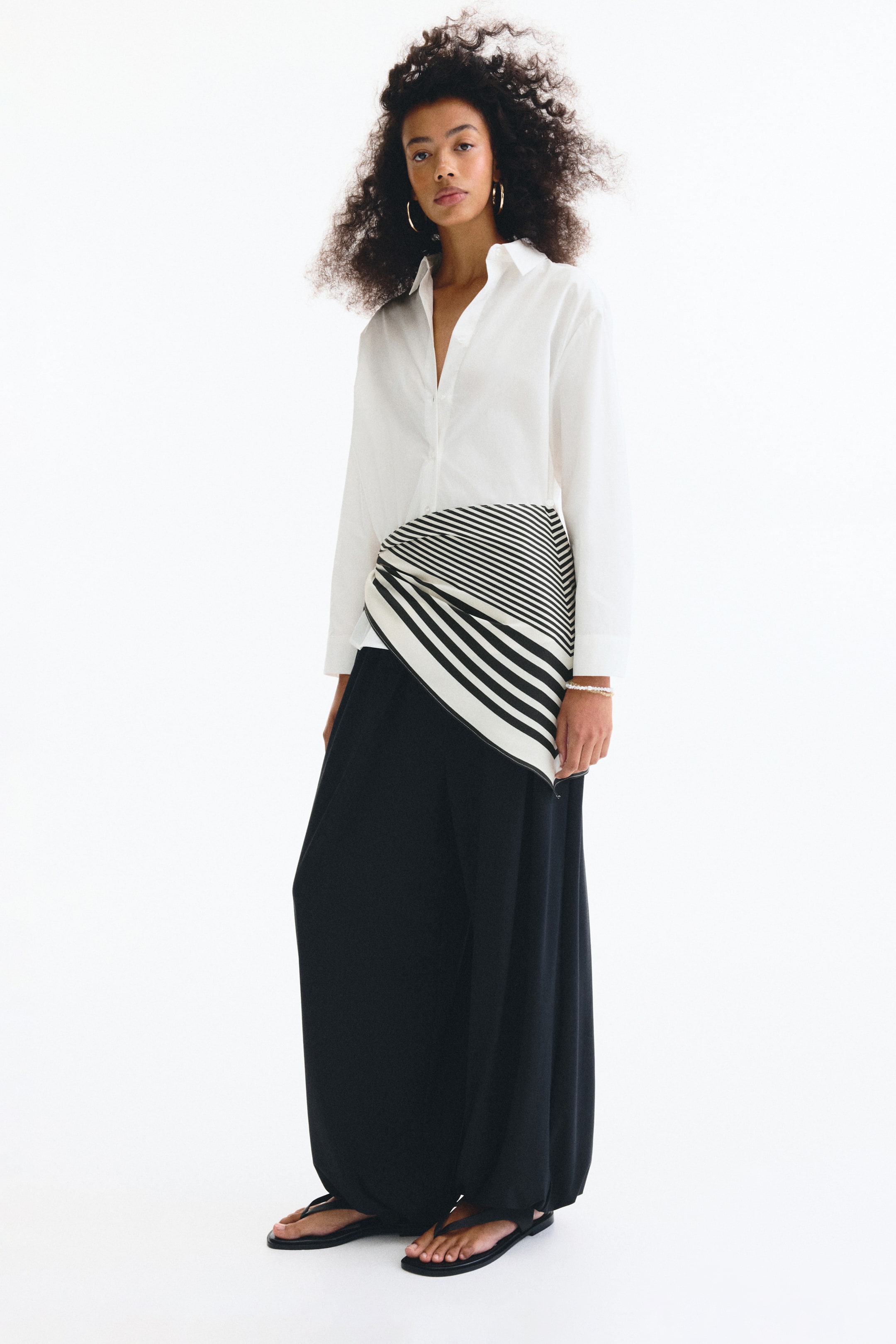 Ampliar la imagen: Se lleva puesta una camisa blanca de manga larga con cuello, superpuesta con una tela de rayas diagonales en blanco y negro envuelta alrededor de la cintura. Unos pantalones negros de pierna ancha, que se recogen en los tobillos, completan el conjunto con sandalias planas negras tipo tanga.