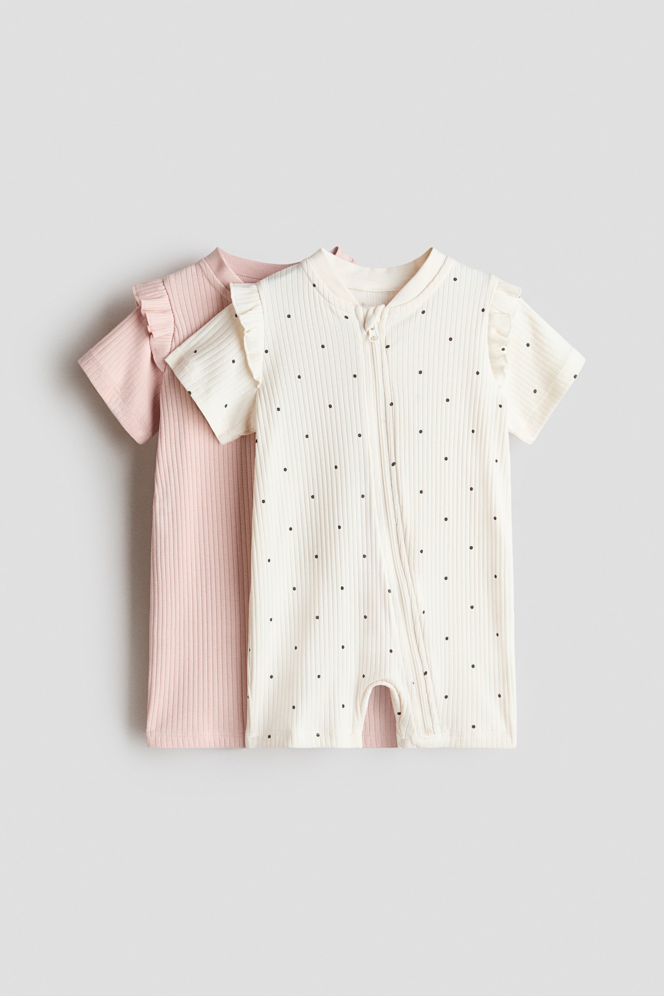 View larger image: A cream-colored baby romper with black polka dots is showcased from the front, featuring short ruffled sleeves, a ribbed texture, and a diagonal front zipper. It is layered over a plain pink baby romper, also featuring short ruffled sleeves and a ribbed texture.