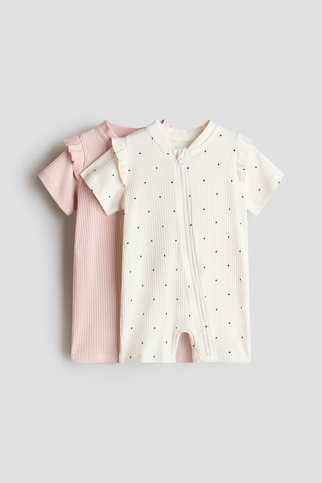 A cream-coloured baby romper with black polka dots is showcased from the front, featuring short ruffled sleeves, a ribbed texture, and a diagonal front zip. It is layered over a plain pink baby romper, also featuring short ruffled sleeves and a ribbed texture. - 1