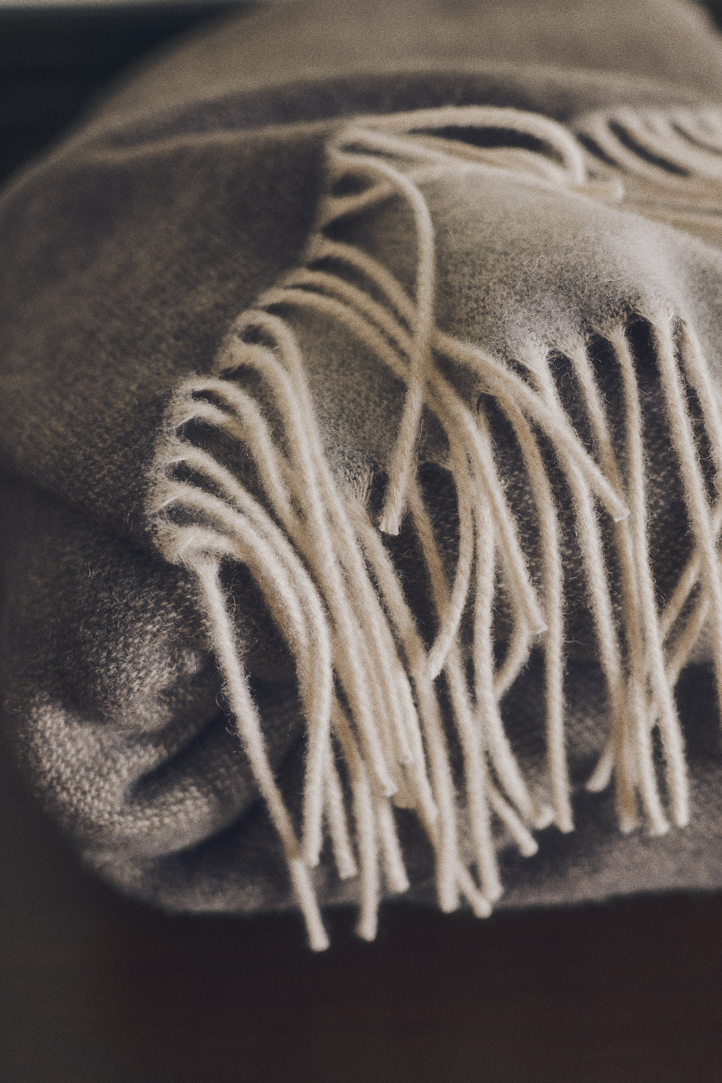 A wool-blend blanket in a textured taupe-brown melange is shown folded from the front, showcasing a soft woven texture and contrasting cream-colored fringes along the bottom edge.