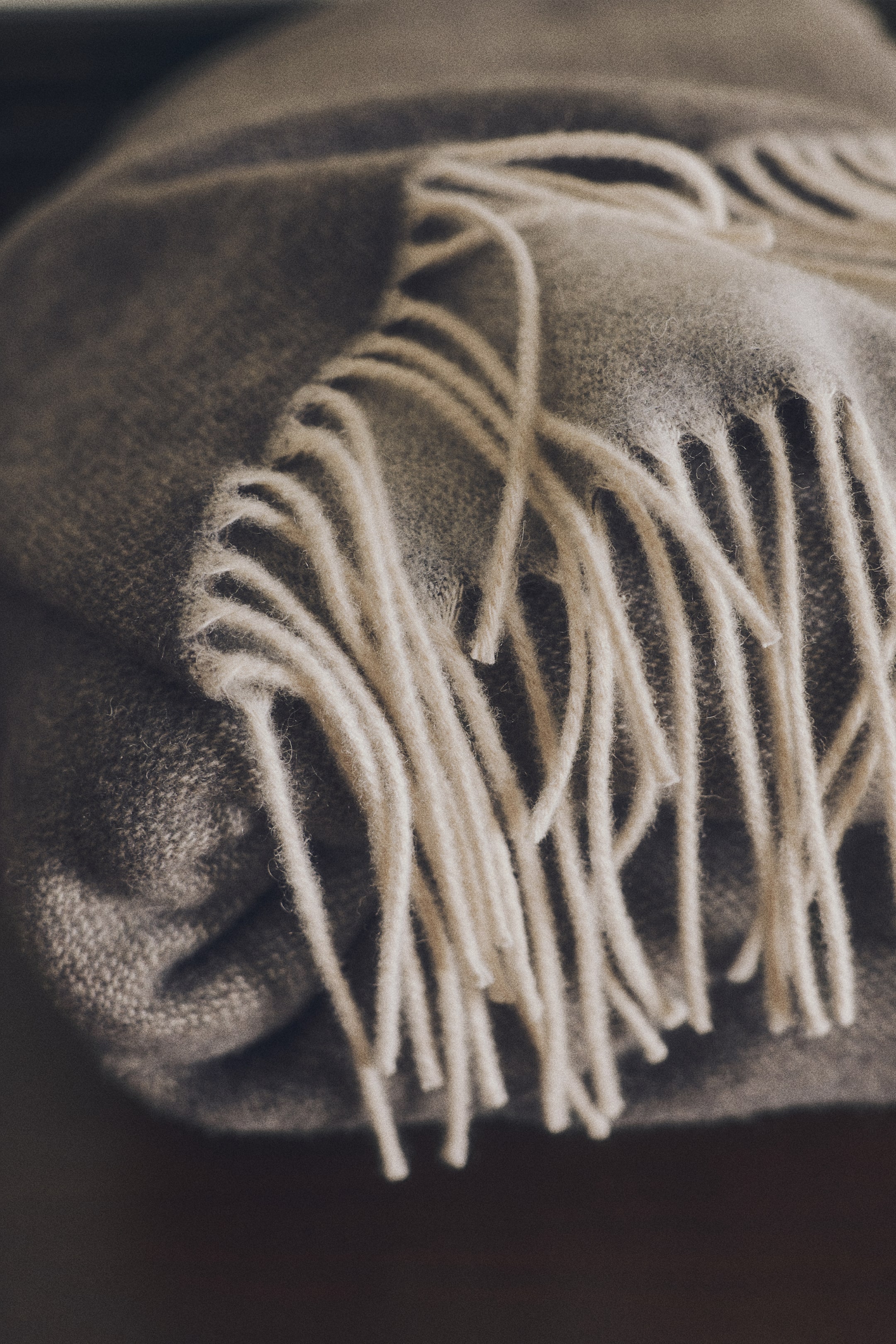 View larger image: A folded wool-blend blanket in a soft taupe shade is showcasing its textured weave and contrasting cream-colored fringe draping down the front.
